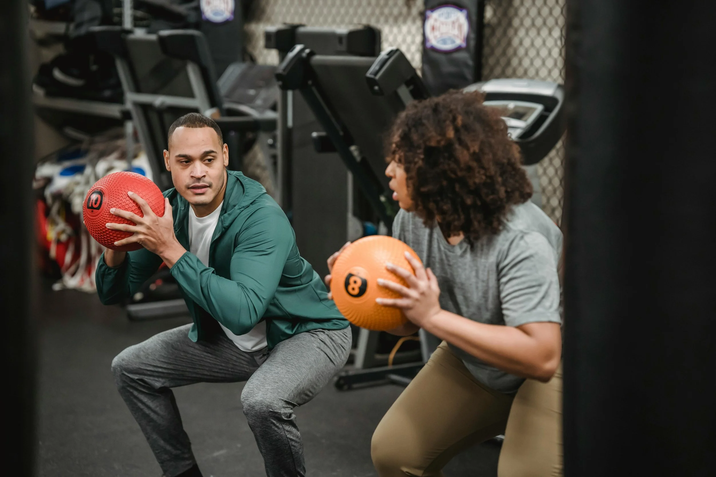 Two people performing squats with medicine balls in a gym. The man on the left has a green jacket and is holding a red medicine ball, while the woman on the right is wearing a gray shirt and beige pants, holding an orange medicine ball.