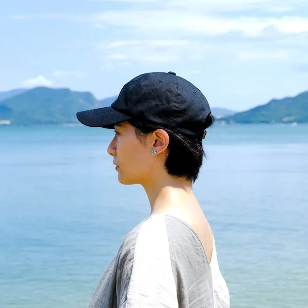 A woman with short hair wearing a black cap, earrings, and a light-colored shirt, standing outdoors by a body of water with mountains in the background.