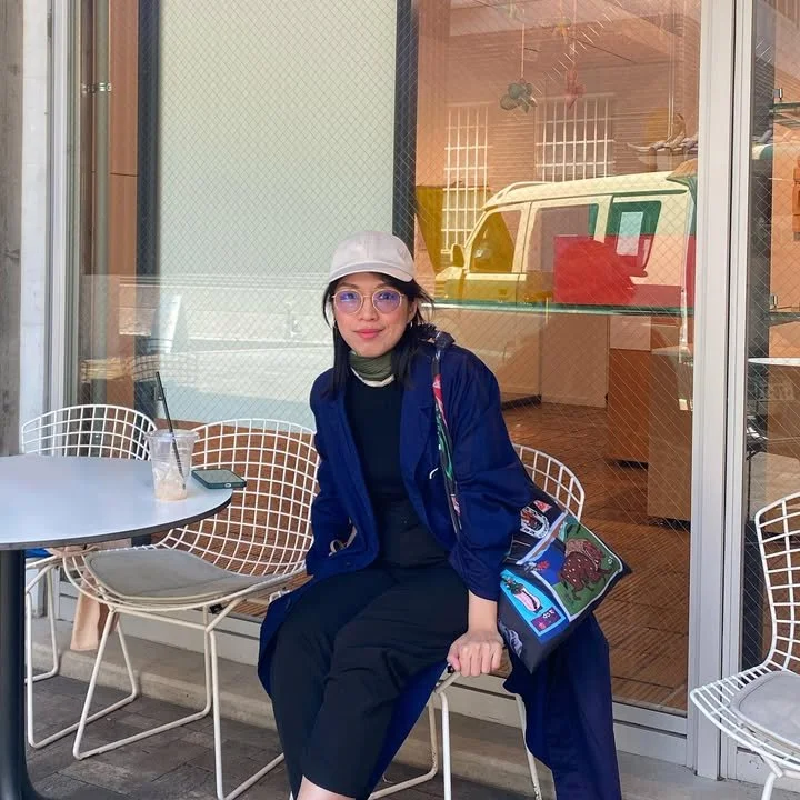 A woman sitting outside a cafe with a white hat, glasses, and a blue coat, holding a colorful bag, next to a table with a drink and a phone.