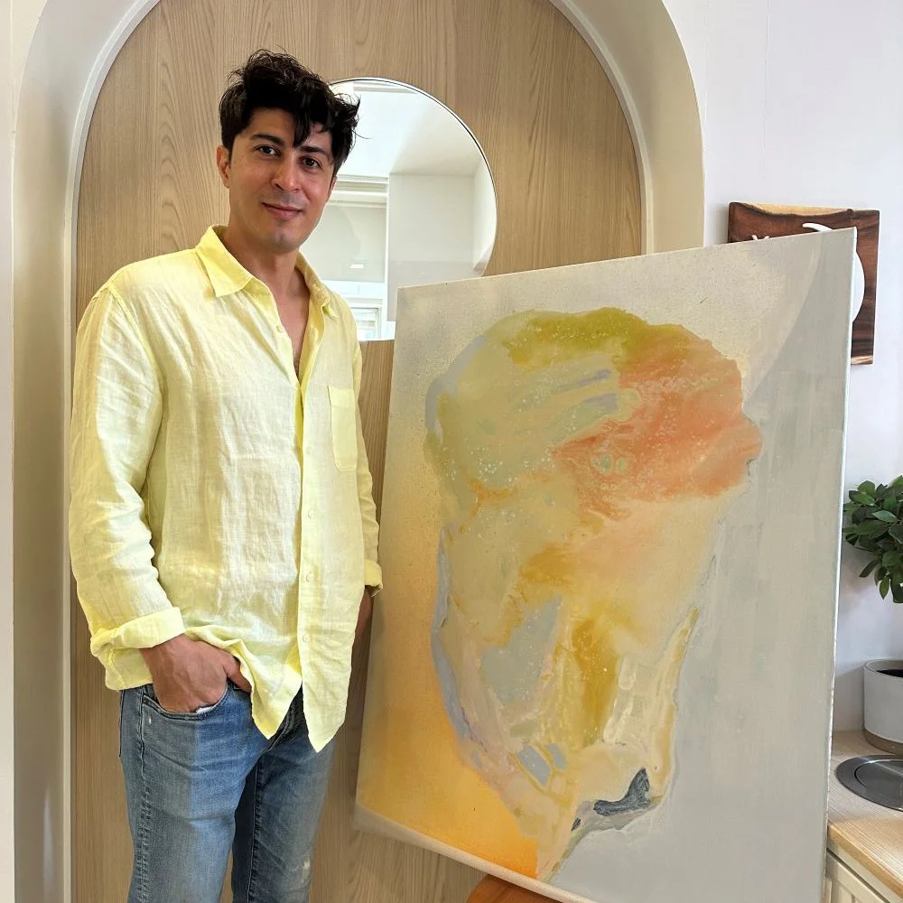 A man with dark hair wearing a yellow shirt and jeans standing next to an abstract watercolor painting of a face in profile.