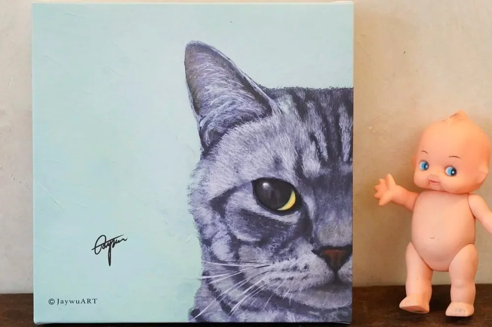 A painting of a gray tabby cat's face on a light blue background, signed by JaywuART, with a plastic doll baby standing next to it on the right side.
