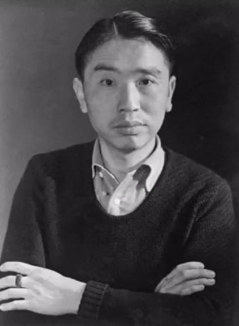 Black and white portrait of a young man with short hair, wearing a collared shirt and a sweater, seated with arms crossed.