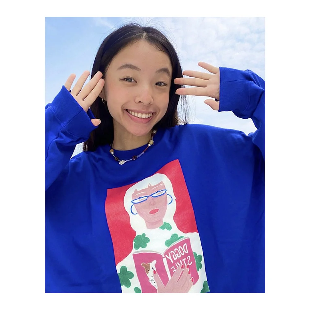 A young woman with long dark hair smiling, wearing a blue sweatshirt with a cartoon image of a woman reading a book titled "Doggy Style" and a beaded necklace, outdoors against a blue sky with some clouds.