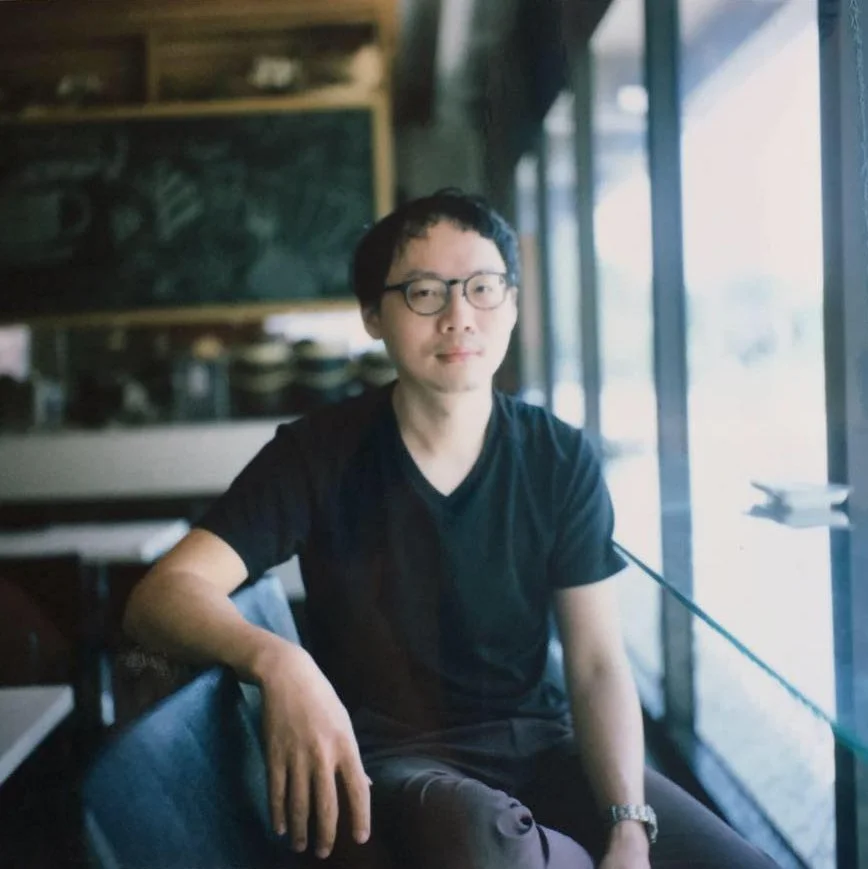 A man with glasses and black hair sitting in a cafe, wearing a black t-shirt, near large windows with sunlight coming through.