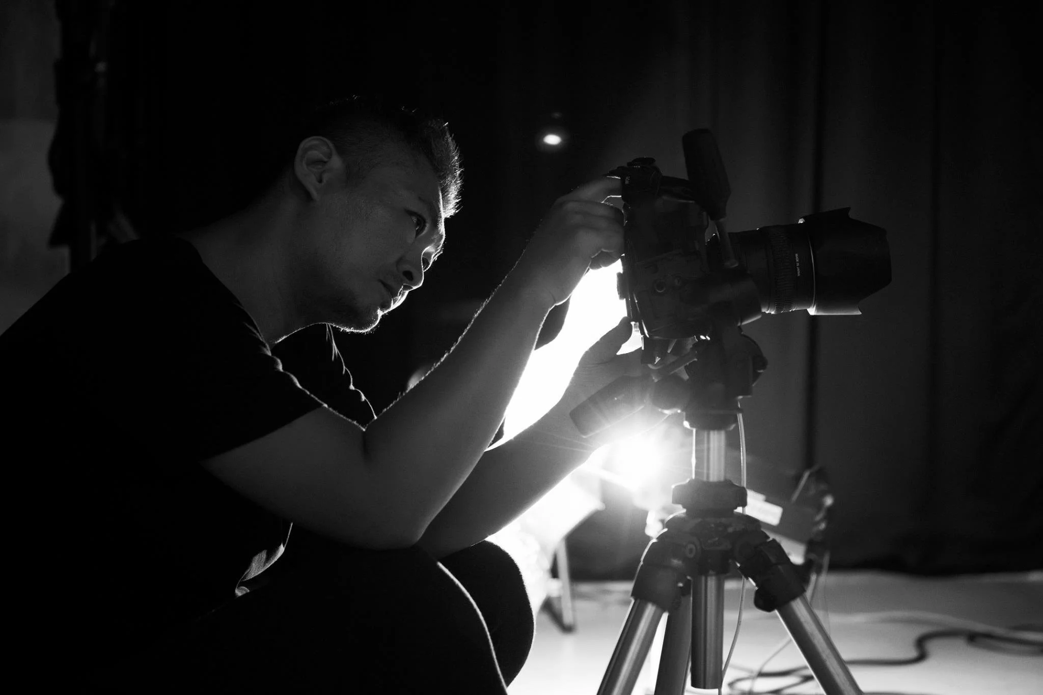 A person setting up or adjusting a professional camera on a tripod in a dark room with backlighting.