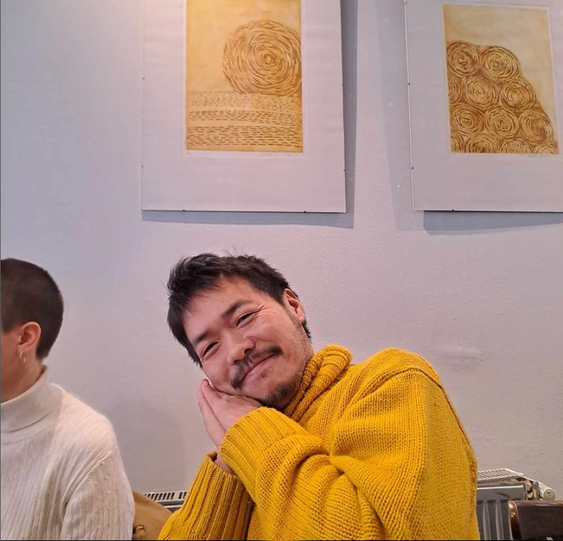 A man with short dark hair and a beard, wearing a yellow sweater, is smiling and resting his head on his hands in a cozy pose. Behind him, two framed artworks are hanging on a white wall, with the left piece featuring a spiral pattern and the right piece depicting swirling circular designs.