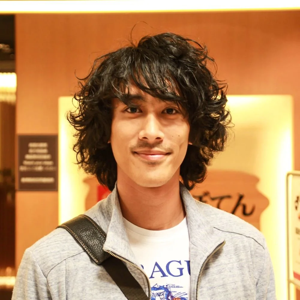 Young man with curly, tousled black hair smiling, wearing a light gray zip-up jacket and a white graphic T-shirt, with a backpack strap over his shoulder, standing indoors in front of a warm-colored wall with a sign in the background.