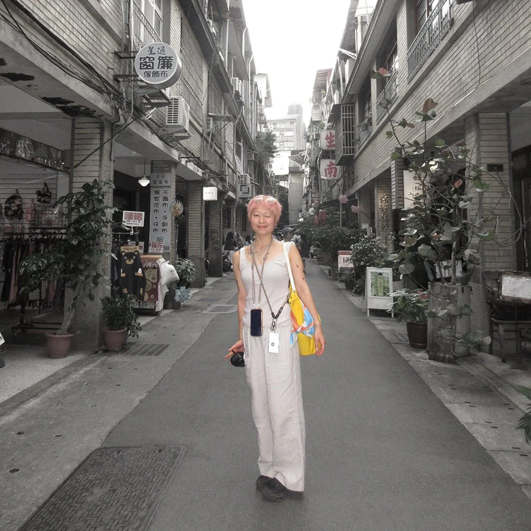 A woman with pink hair wearing white pants and a white top standing in the middle of a narrow city street lined with shops and plants.
