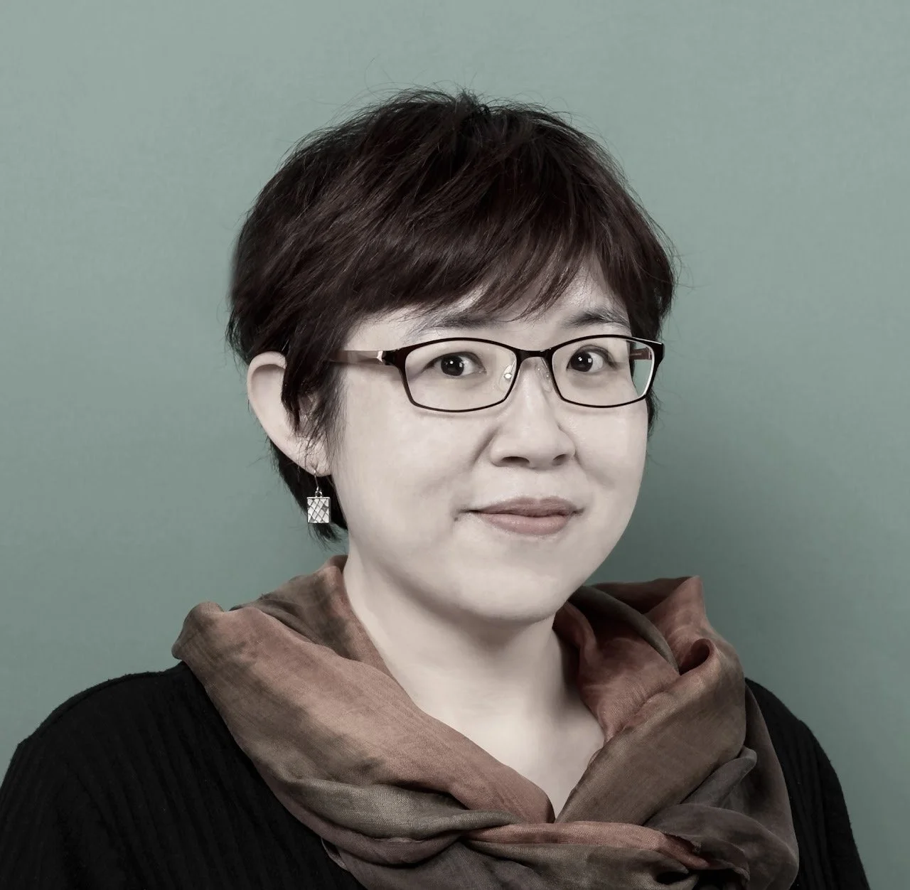 Portrait of a woman with short dark hair, glasses, and earrings, wearing a brown scarf and a black top, against a green background.