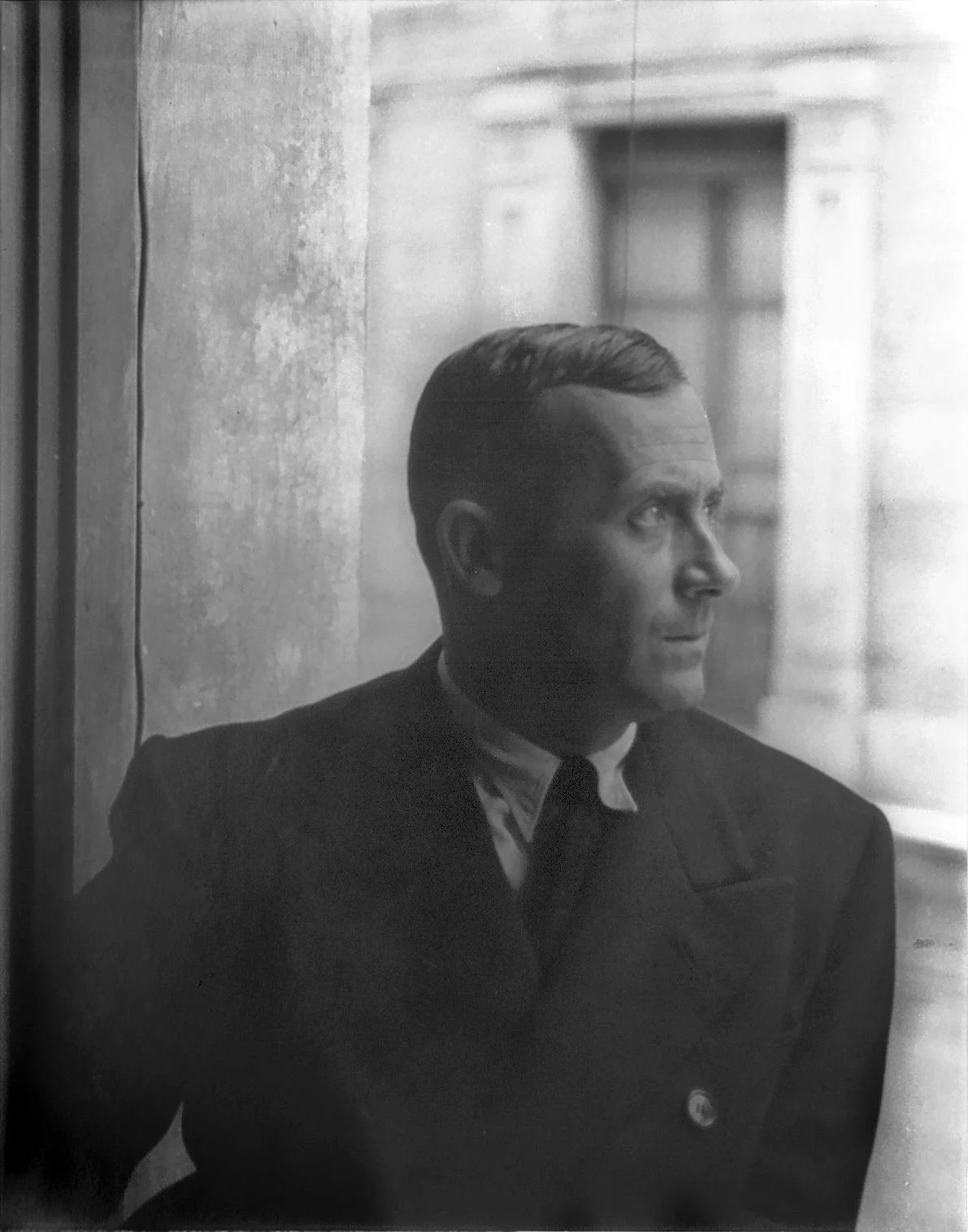 A black and white photograph of a man looking out a window, dressed in a uniform, with a serious expression.