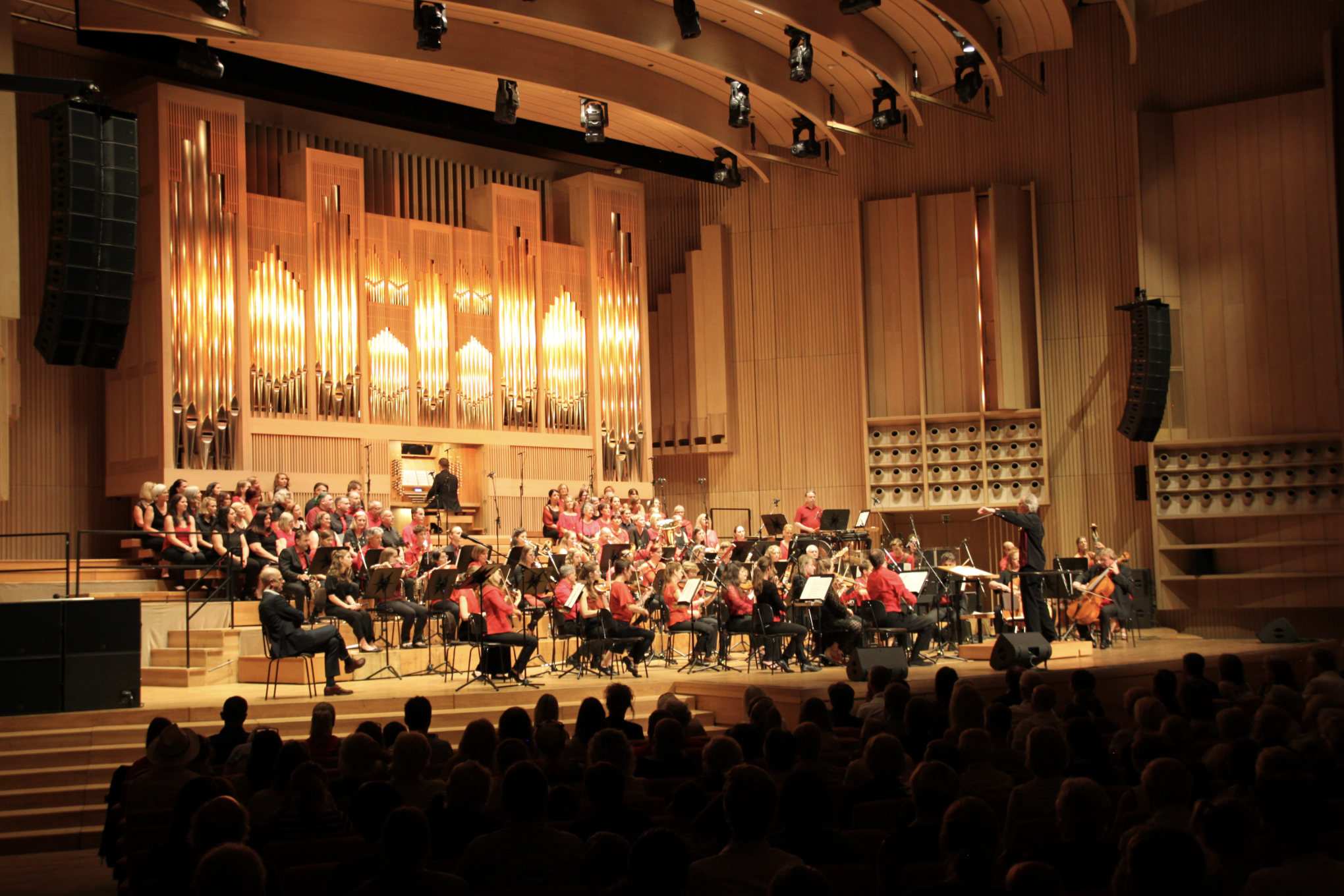 Ein Orchester auf einer Bühne während eines Konzerts, mit einem Dirigenten vorne und einem Publikum im Vordergrund.