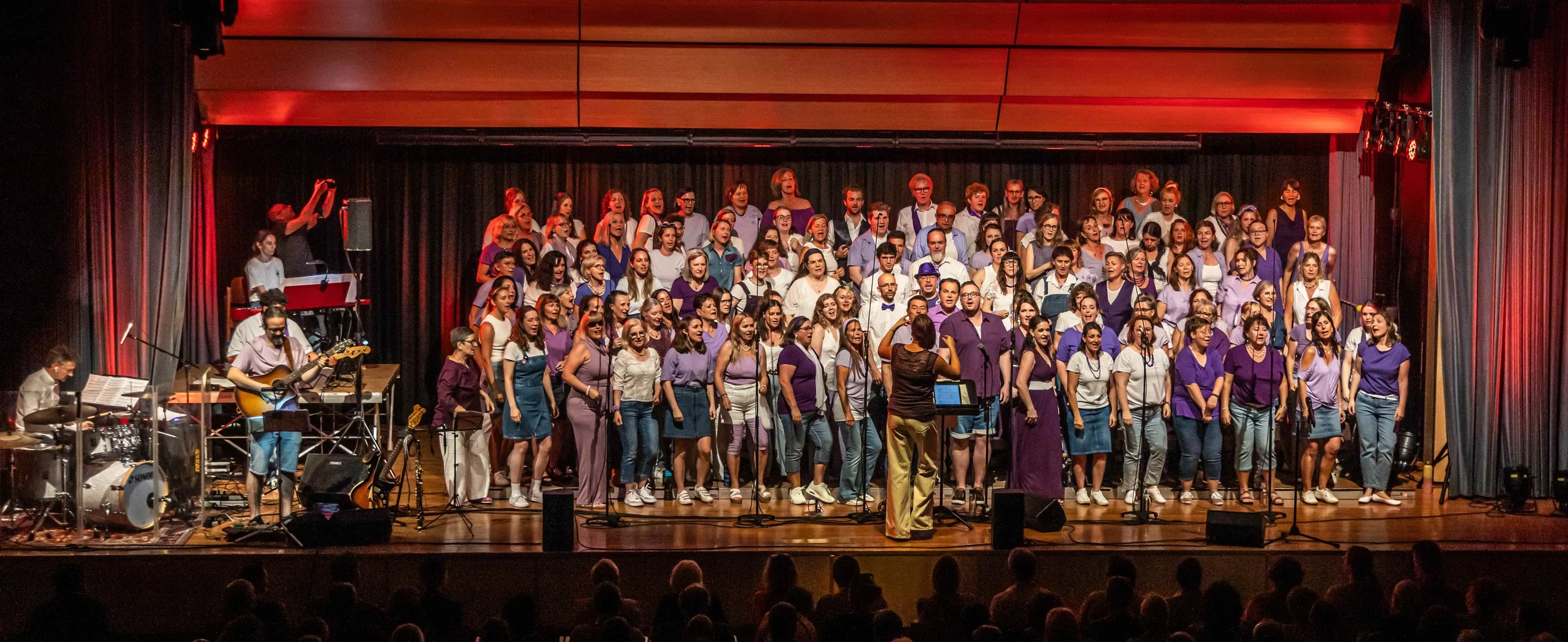 Chor mit Männern und Frauen singt auf einer Bühne, begleitet von einer Band. Die Sänger tragen meist lila, weiß und blaue Kleidung. Es gibt einen Dirigenten und Musiker mit Instrumenten wie Gitarre, Schlagzeug, Keyboard. Im Hintergrund schwarze Vorhä
