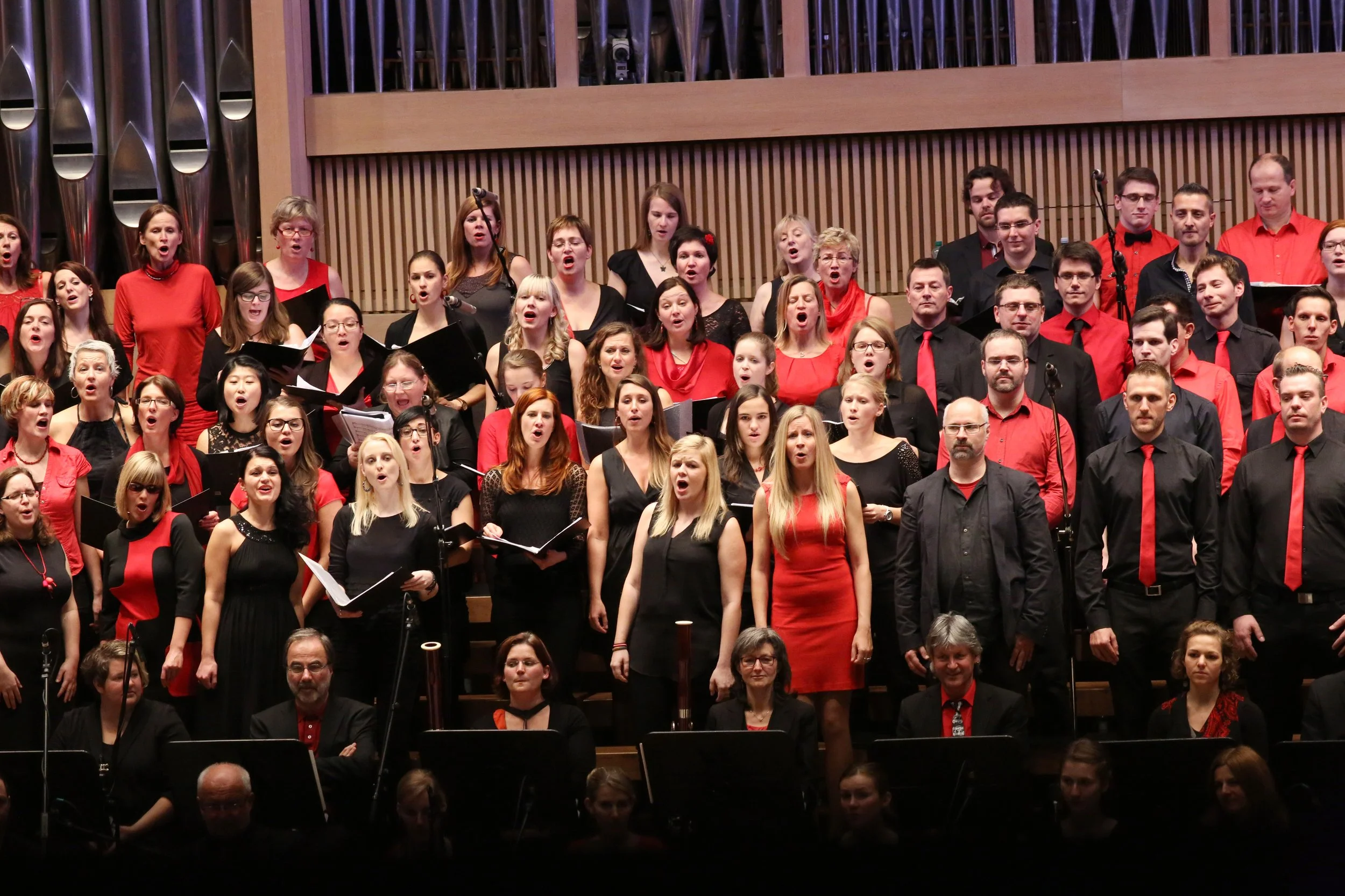 Ein Männer- und Frauenchor performing auf einer Bühne in einem Konzertsaal, viele Sänger tragen schwarz und rot, einige halten Noten, einige singen offen, hinter ihnen eine Holzvertäfelung.