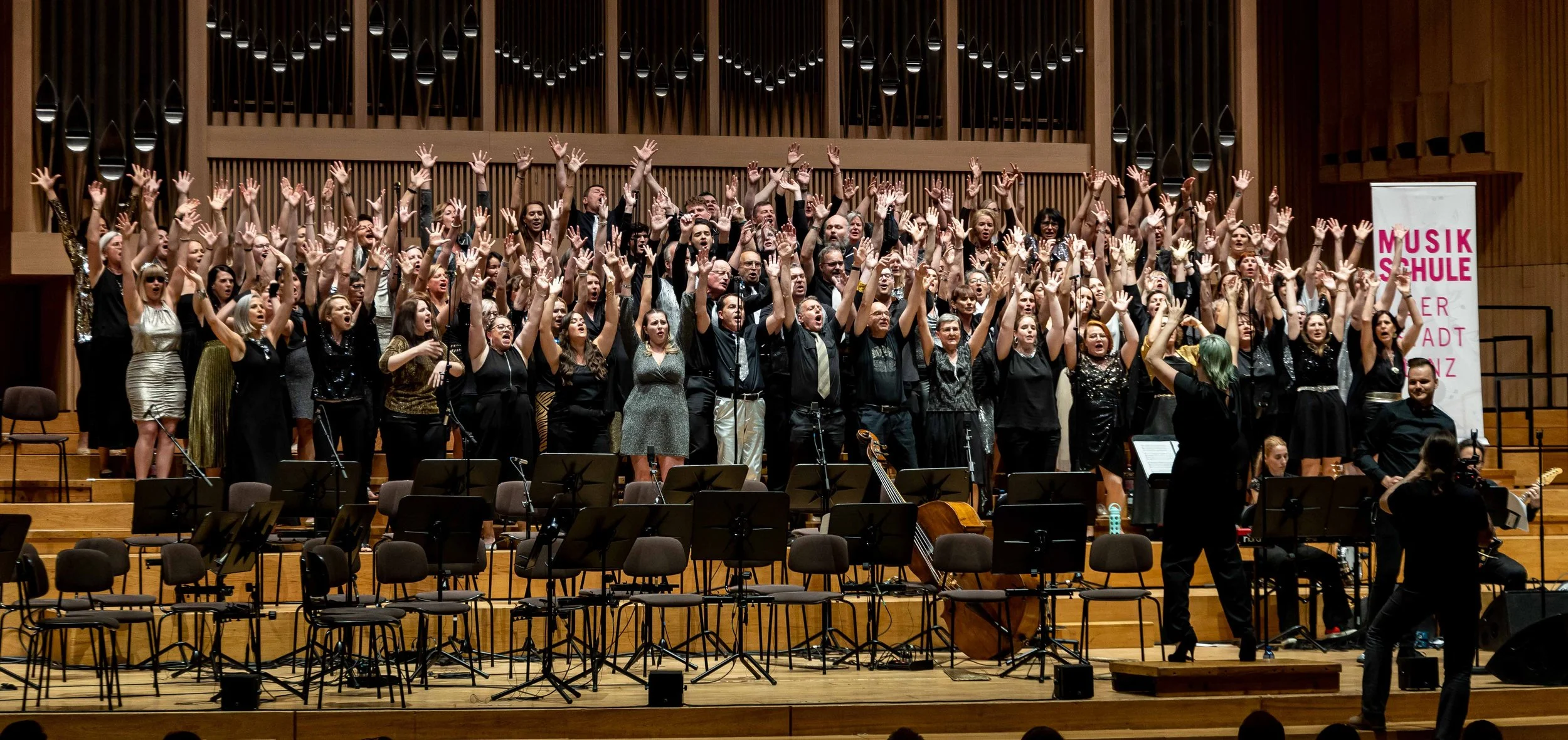 Chor auf einer Bühne mit Musikern, Dirigent, Klavier, Gitarren und Sitzgelegenheiten, während sie singen.
