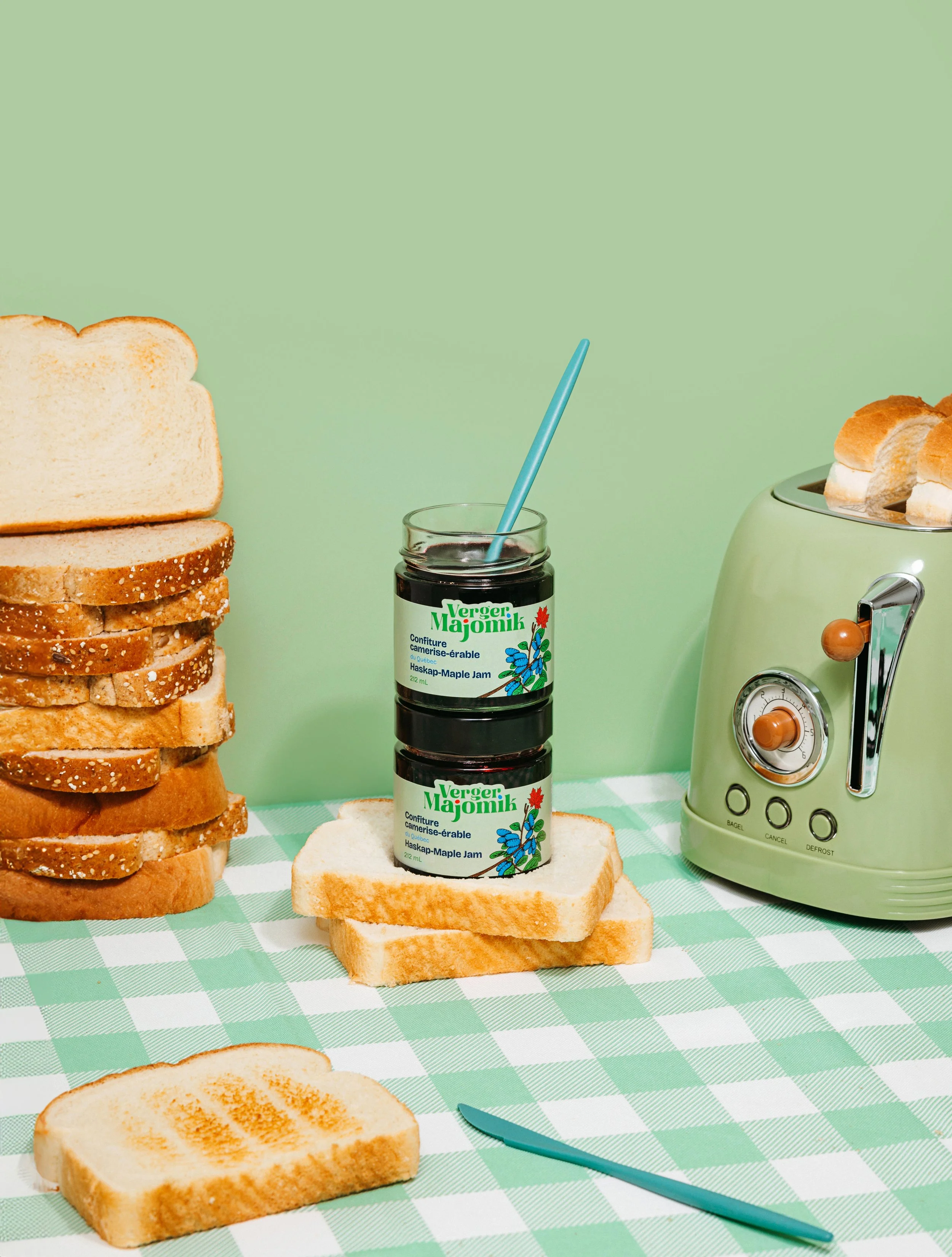 Stacks of sliced white bread, jars of maple jam, a toaster with slices of bread, and a checkered green and white tablecloth.