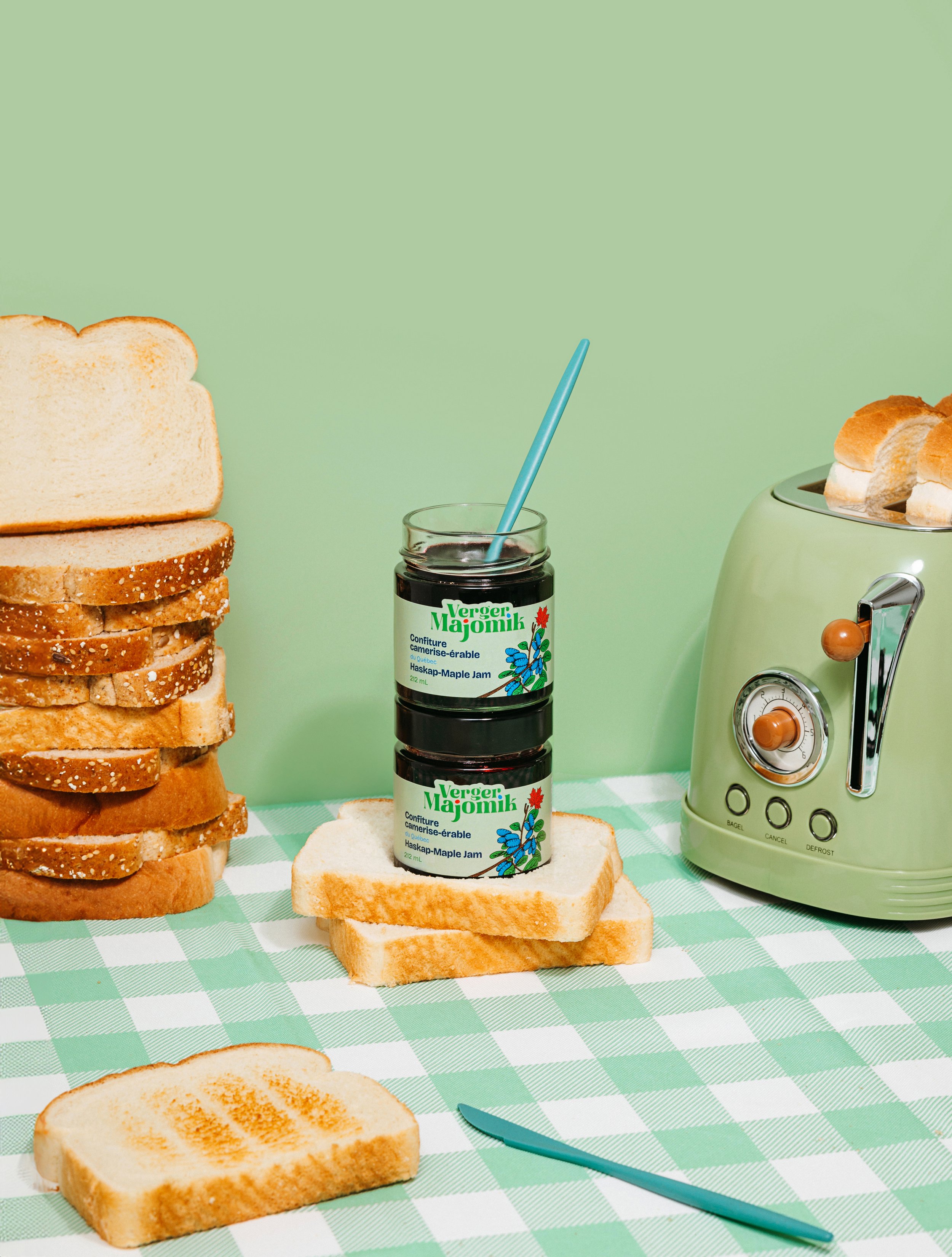 Stacks of sliced white bread, jars of maple jam, a toaster with slices of bread, and a checkered green and white tablecloth.