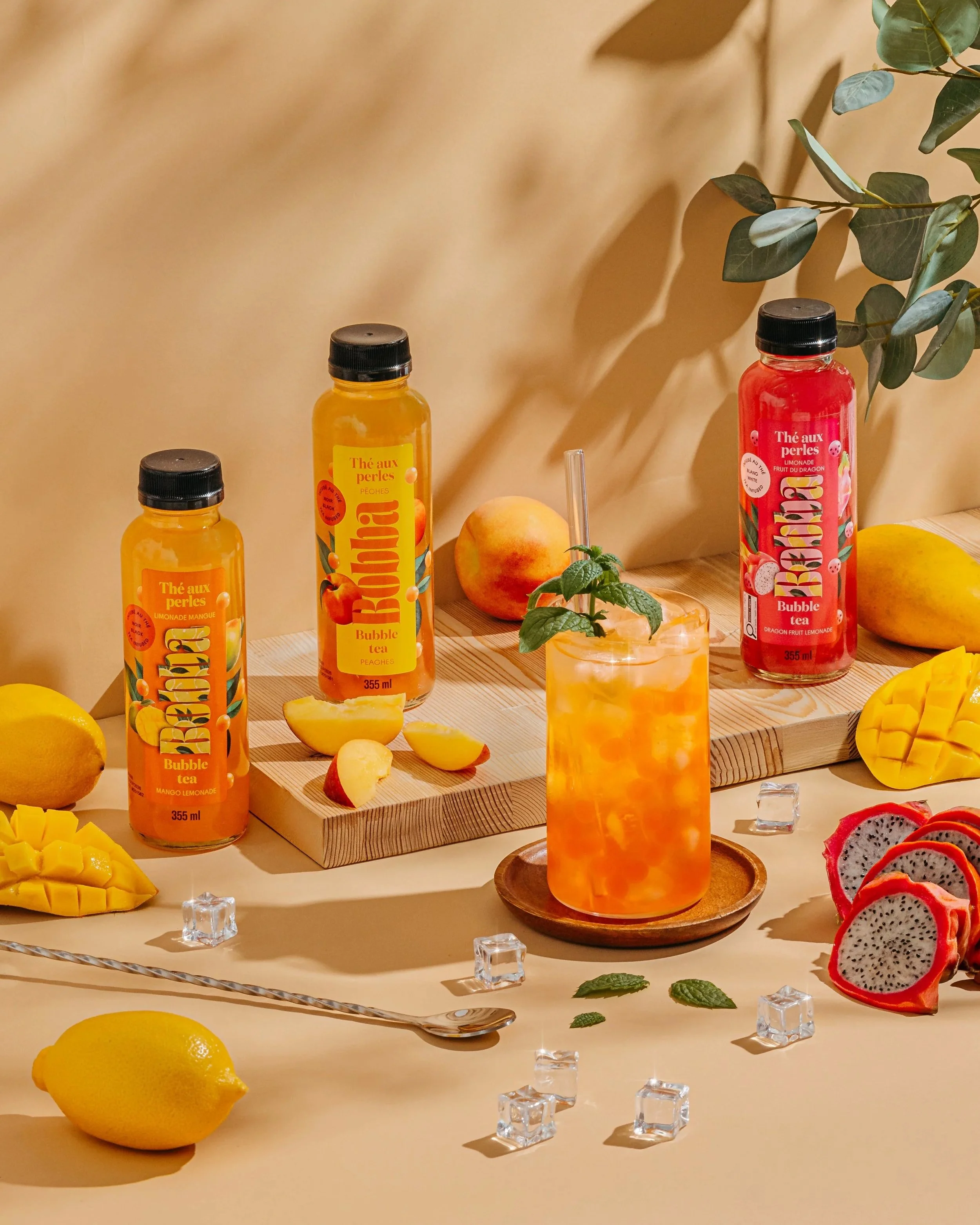 Display of bottled fruit flavored teas, including peach and berry flavors, surrounded by fresh fruits such as lemon, peach, mango, and dragon fruit. The setup includes a light-colored wooden surface, a beige background with a shadow of leaves, and some decorative elements like mint leaves and ice cubes.
