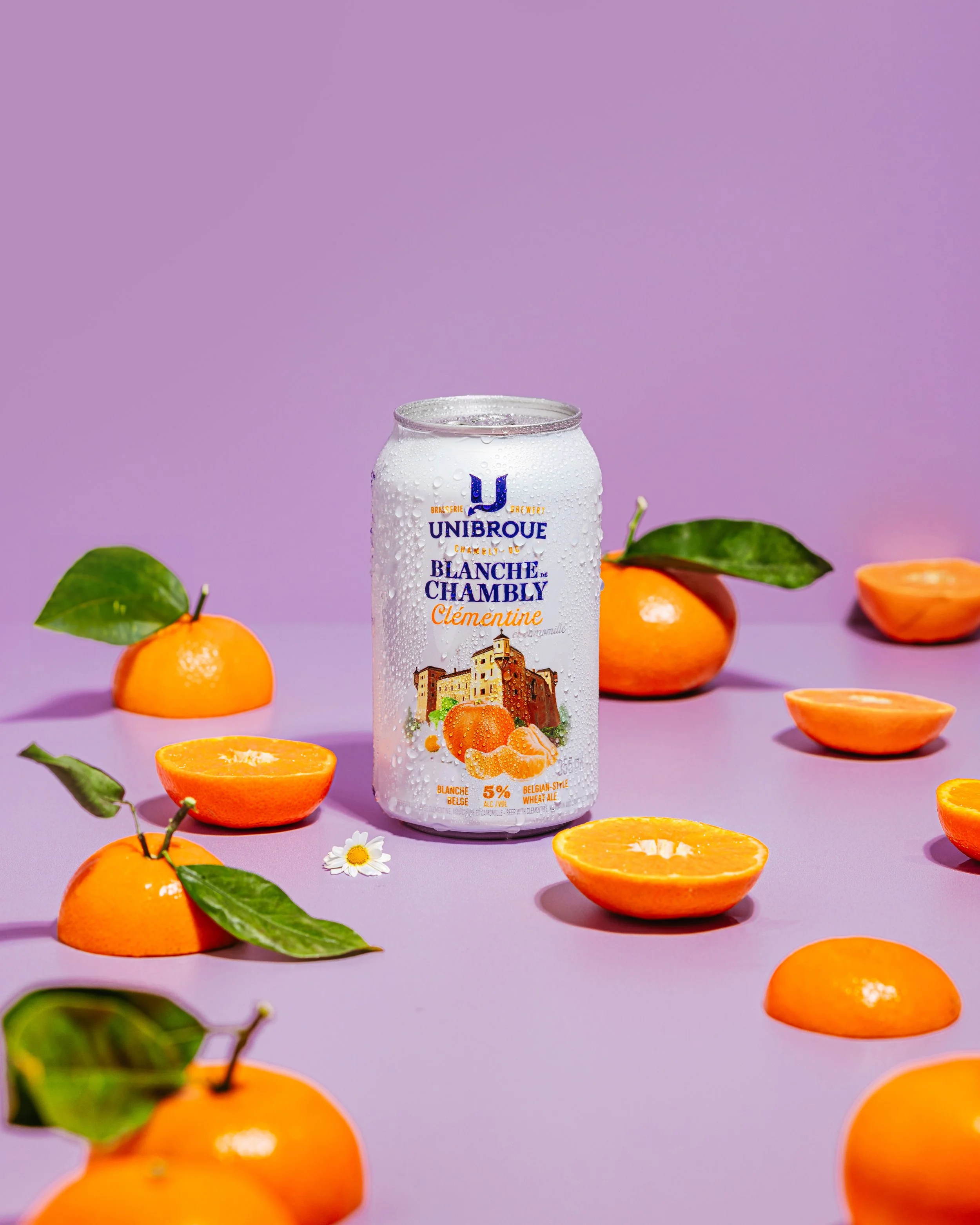 A can of blanc de Chambly Clémentine soda surrounded by whole and halved clementines with green leaves, set against a purple background.
