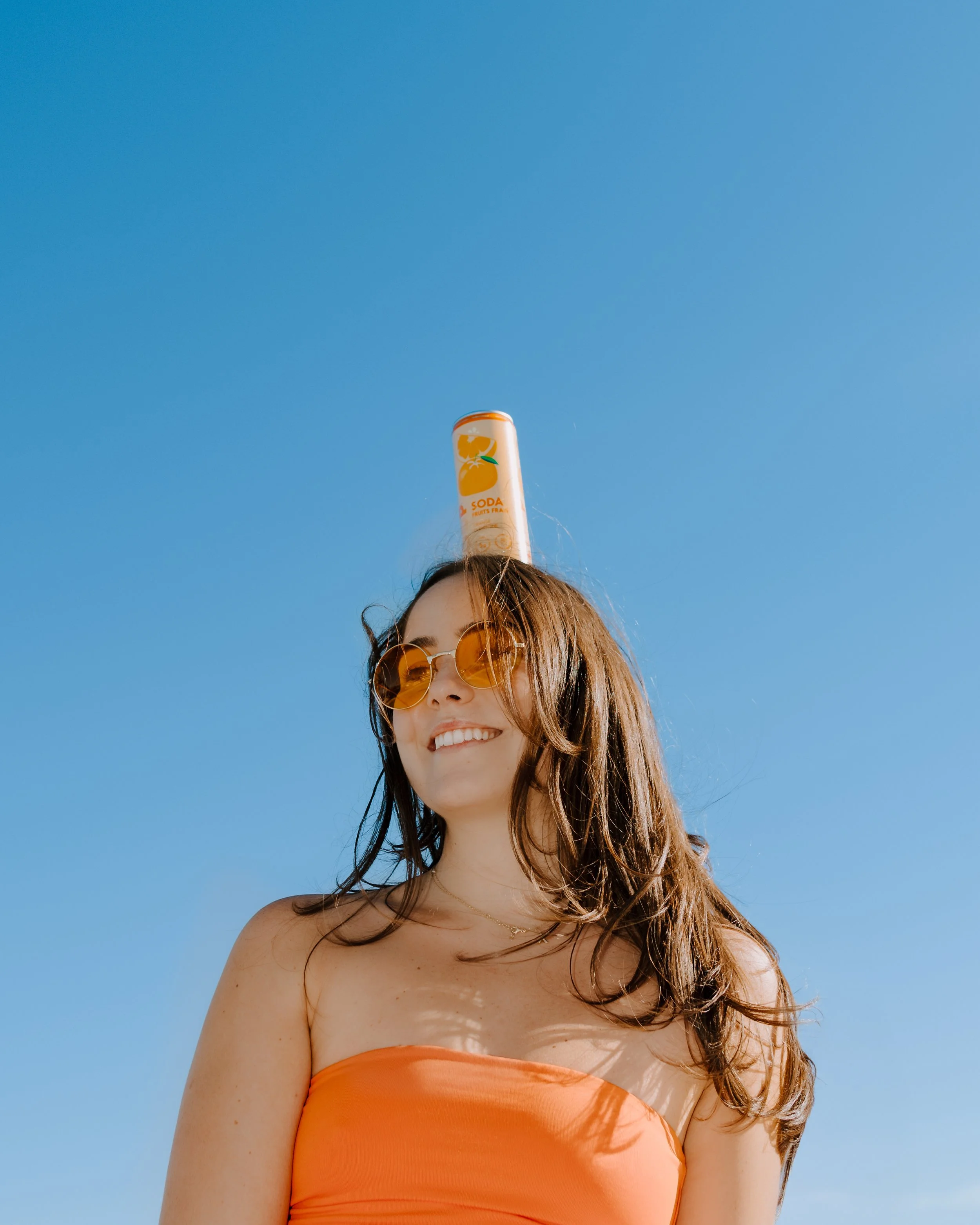 A smiling woman with brown hair wearing orange sunglasses and an orange strapless top, balancing a can of soda on her head against a clear blue sky.