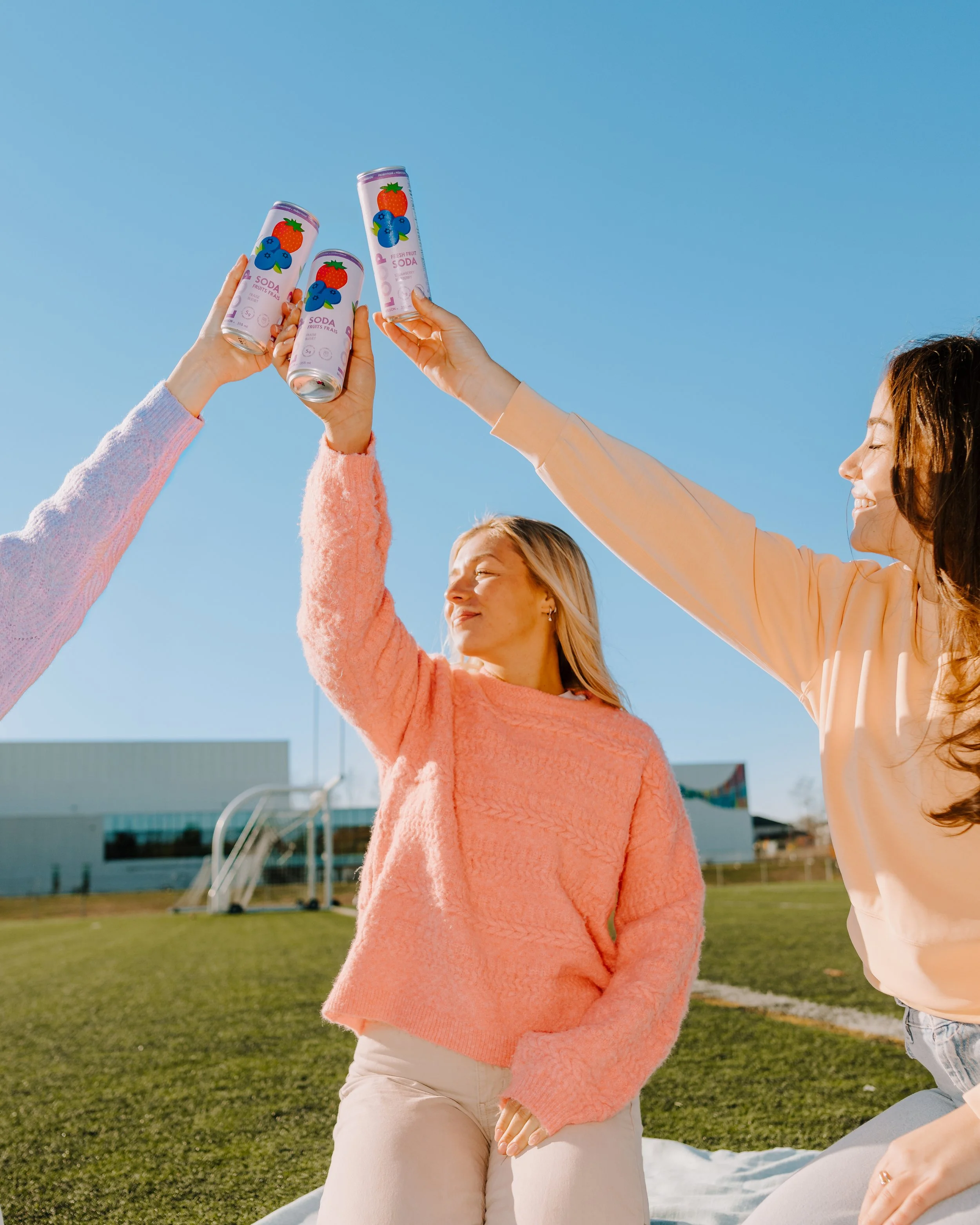 Three women outdoors holding cans of sparkling water, raising them together in a toast against a clear blue sky, with a building and grassy field in the background.