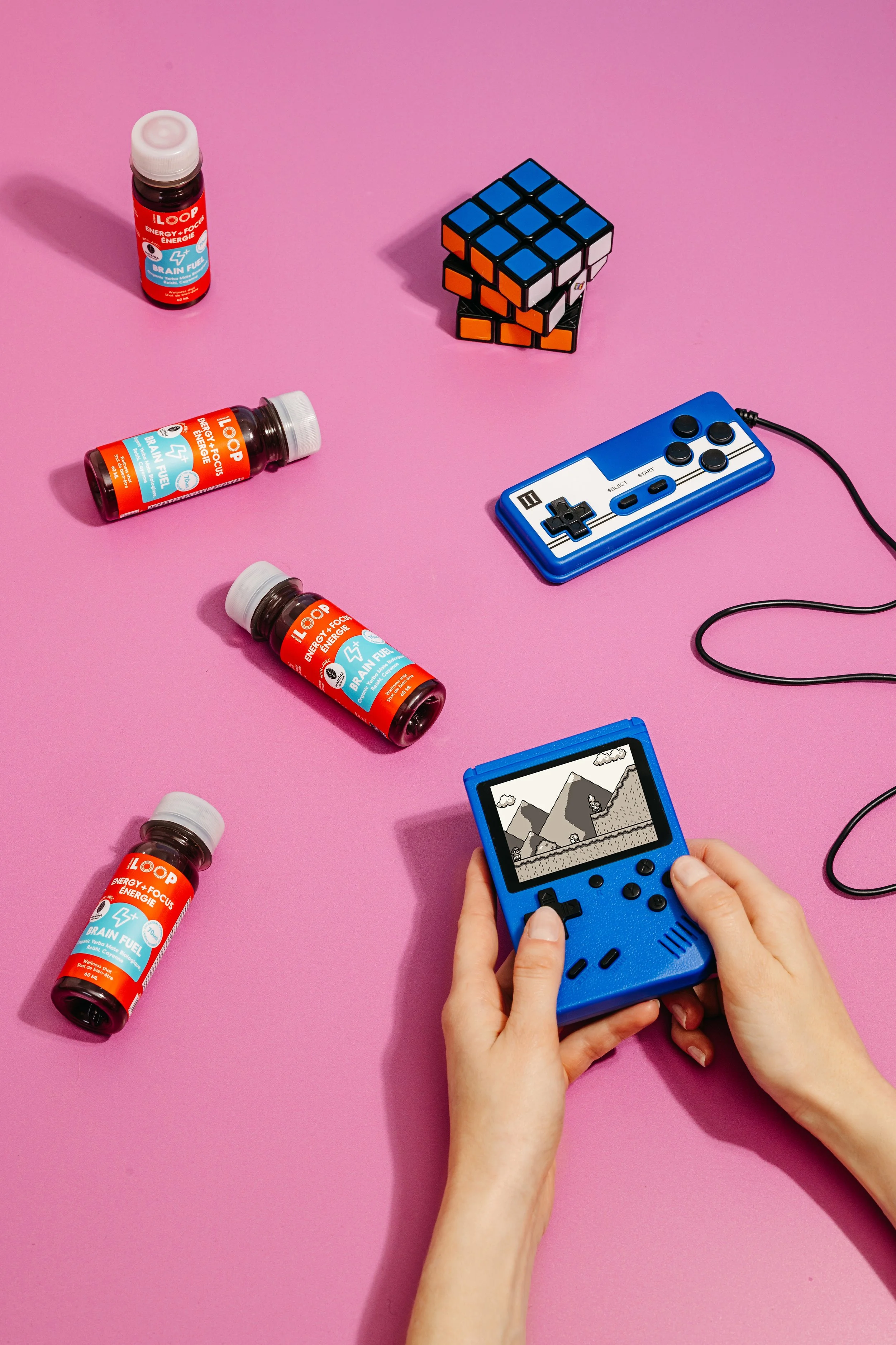 A person's hands holding a blue handheld gaming console playing a pixelated game on a pink surface. Surrounding objects include a small blue and white NES-style gaming controller, a stack of four multi-colored cubed puzzles, a small bottle of energy shots, and another bottle labeled 'Brain Fuel' with a red cap.