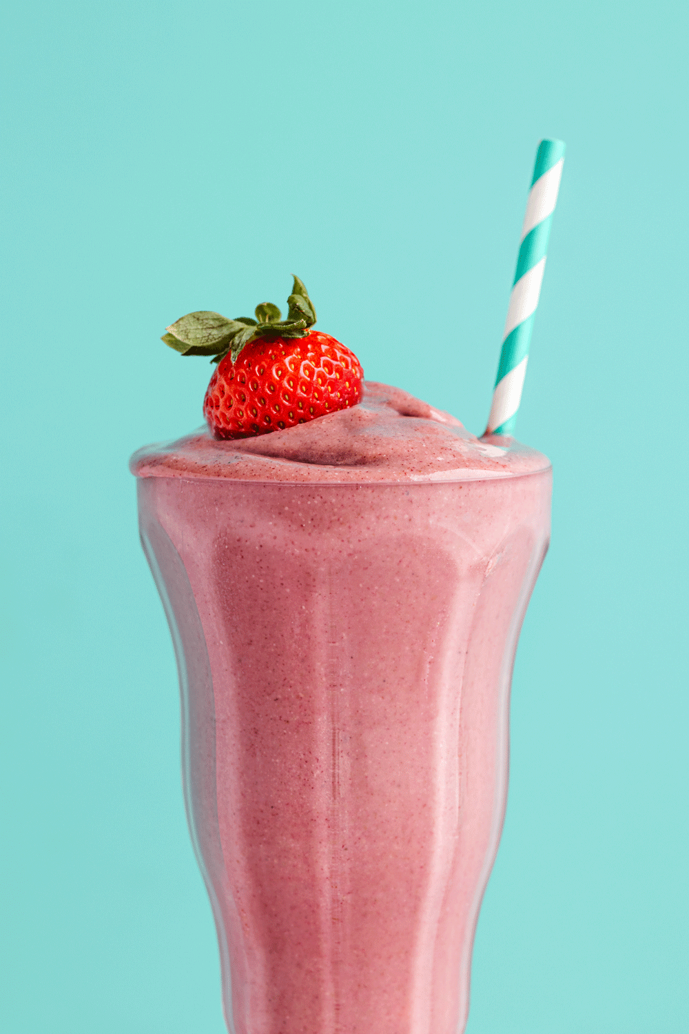 Strawberry smoothie in a tall glass with a strawberry and a striped straw on a light blue background.