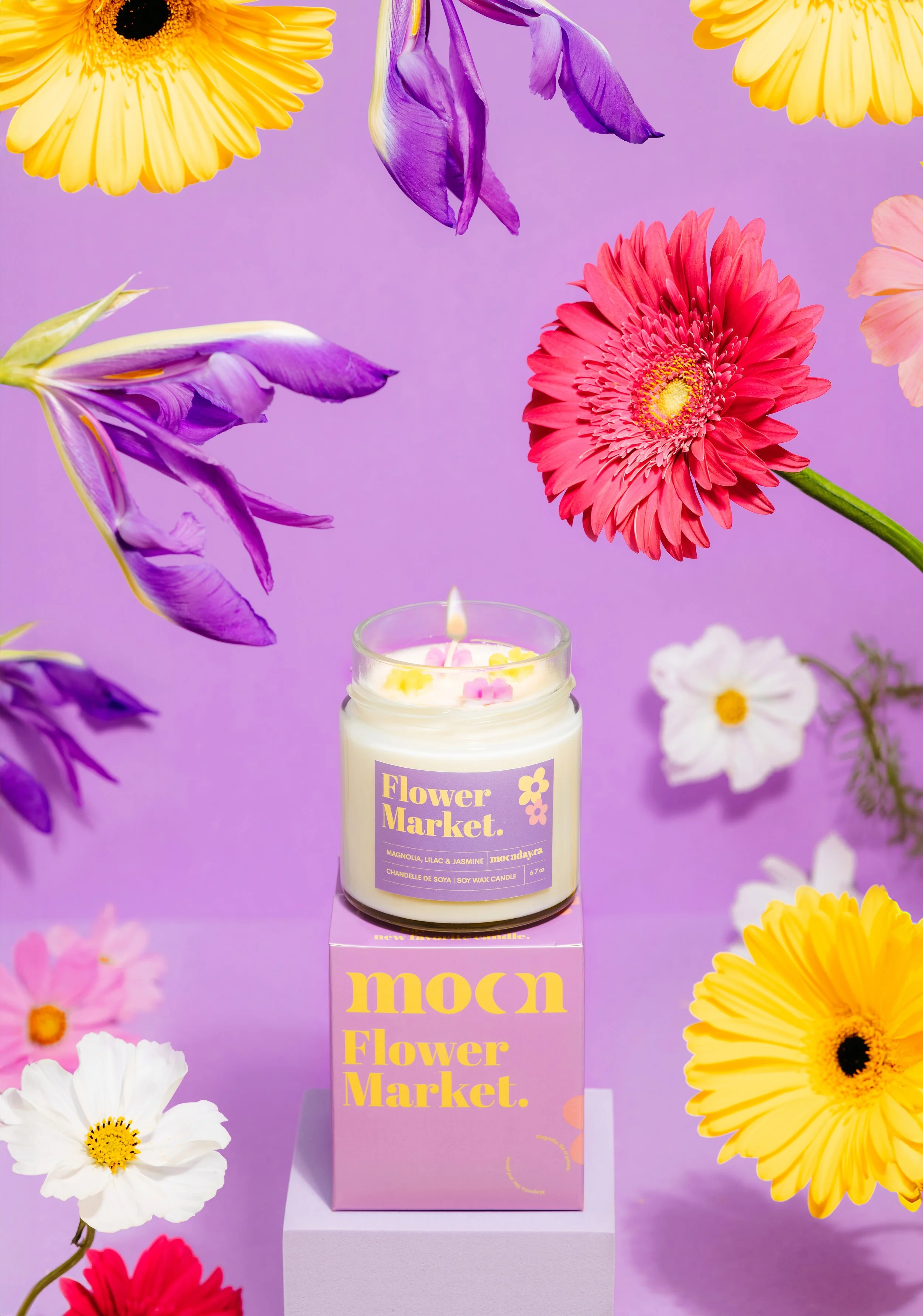 A white candle labeled "Flower Market" with a purple background, surrounded by colorful flowers like yellow daisies, pink gerbera daisies, purple irises, and white cosmos, arranged with some flowers floating and some placed around the candle.