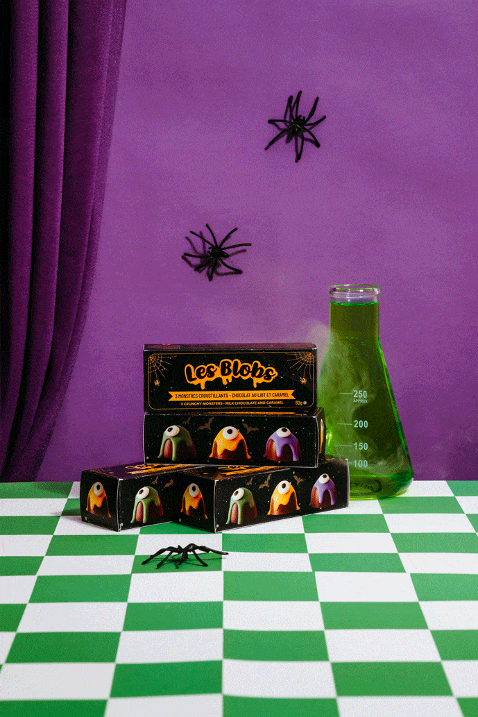 A purple wall with two black plastic spiders hanging, a green liquid-filled measuring beaker, and a black box of chocolate candies with monster faces, placed on a green and white checkered surface.
