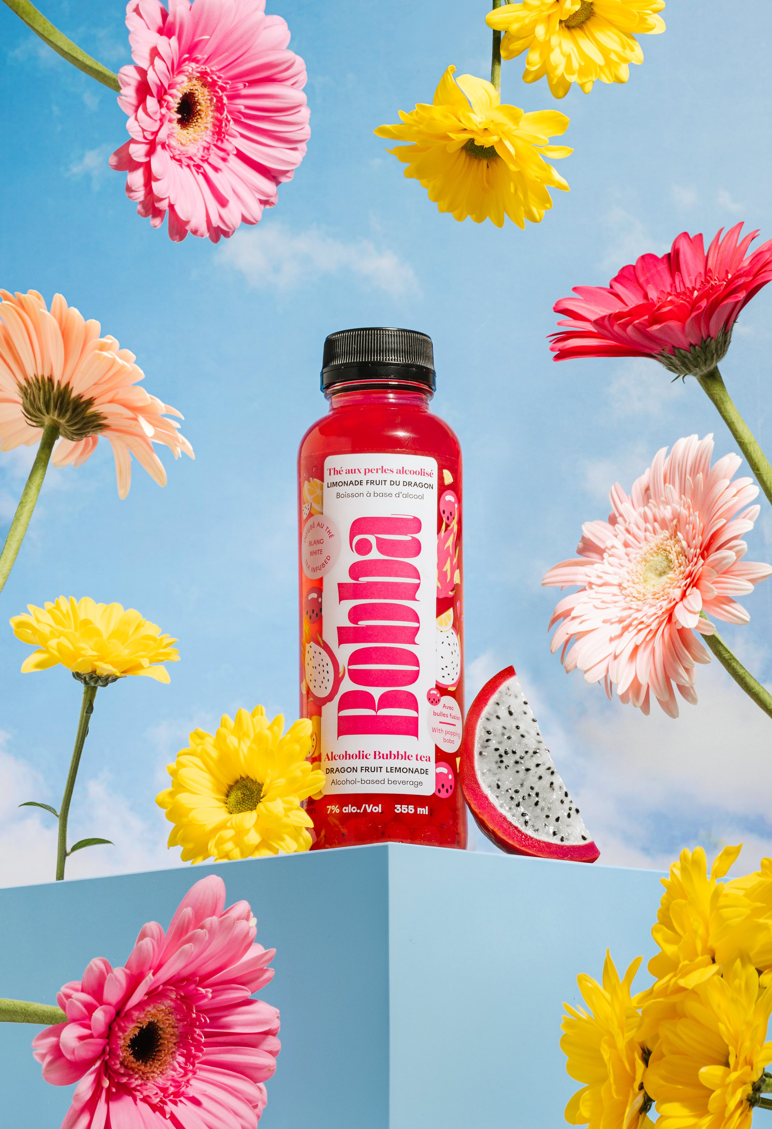 A red bottle of Bubble alcohol bubble tea with dragon fruit lemonade flavor is placed on a light blue surface, surrounded by colorful pink, yellow, and peach daisies, against a bright blue sky background.