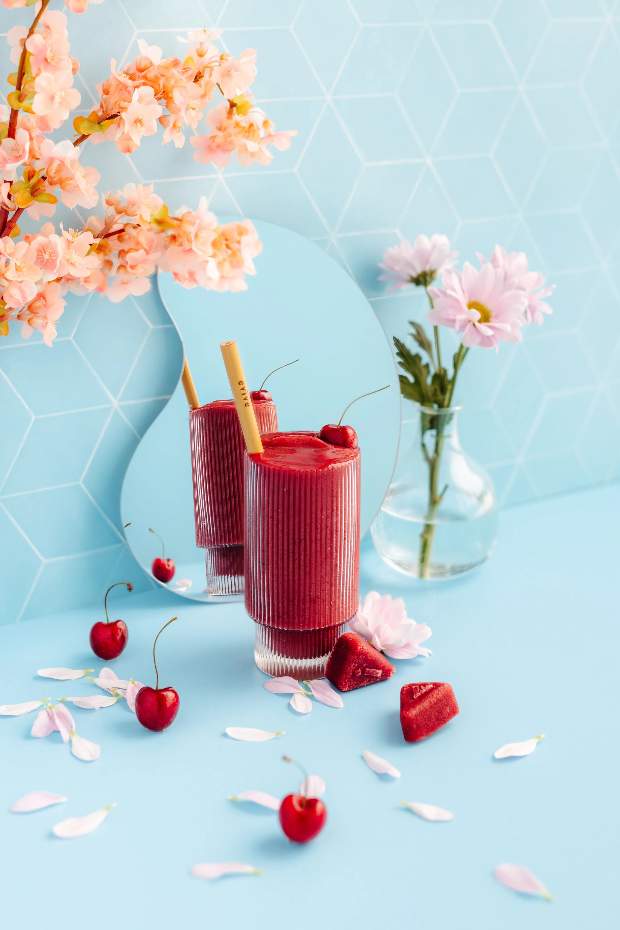 Two pink cherry smoothies with cherries on top, surrounded by petals and cherries, reflected in a round mirror, with pink flowers in a glass vase on a light blue background and blue hexagonal tiled wall.
