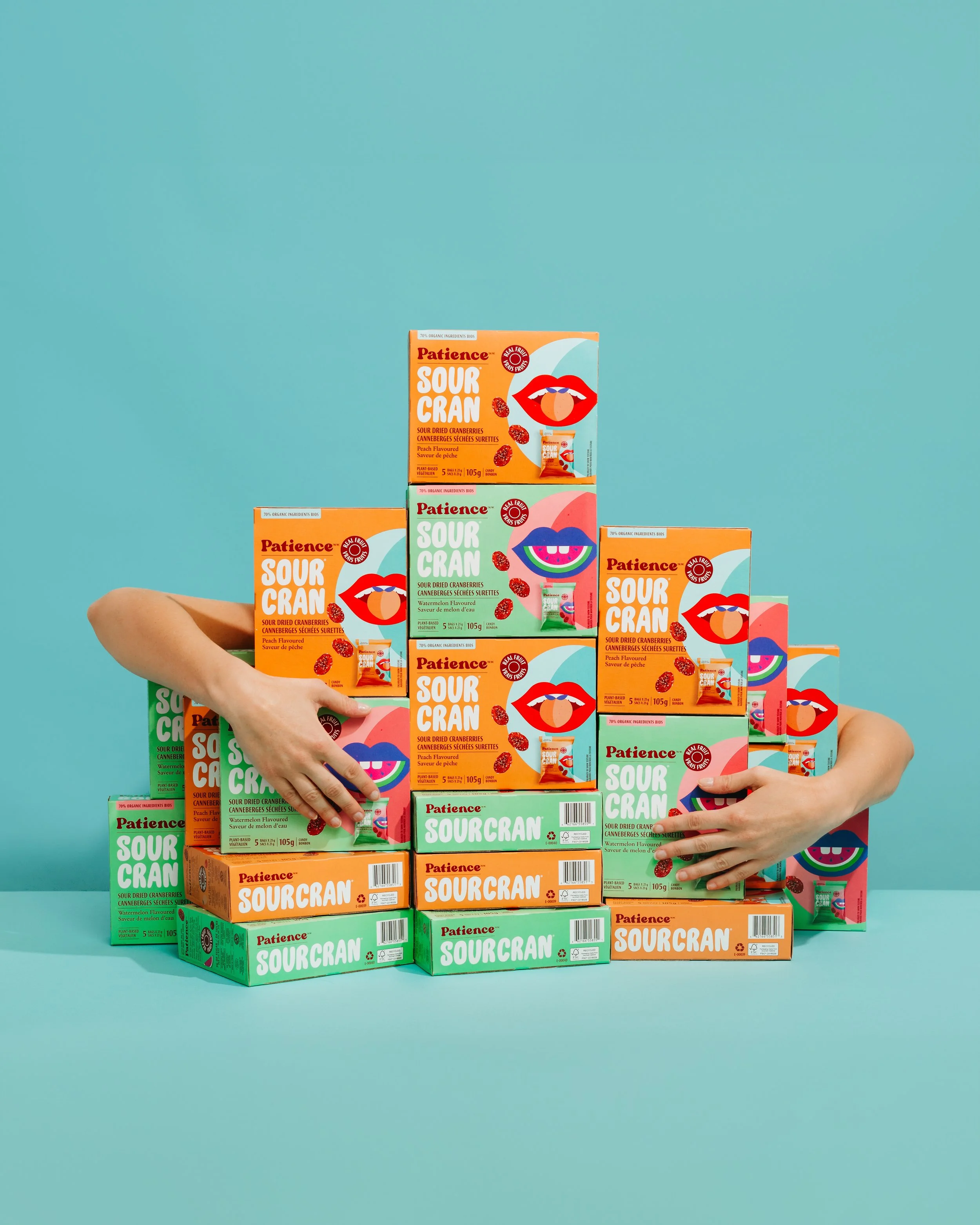 Stack of colorful sour cranberry snack boxes on a bright teal background with two hands reaching in from the sides.