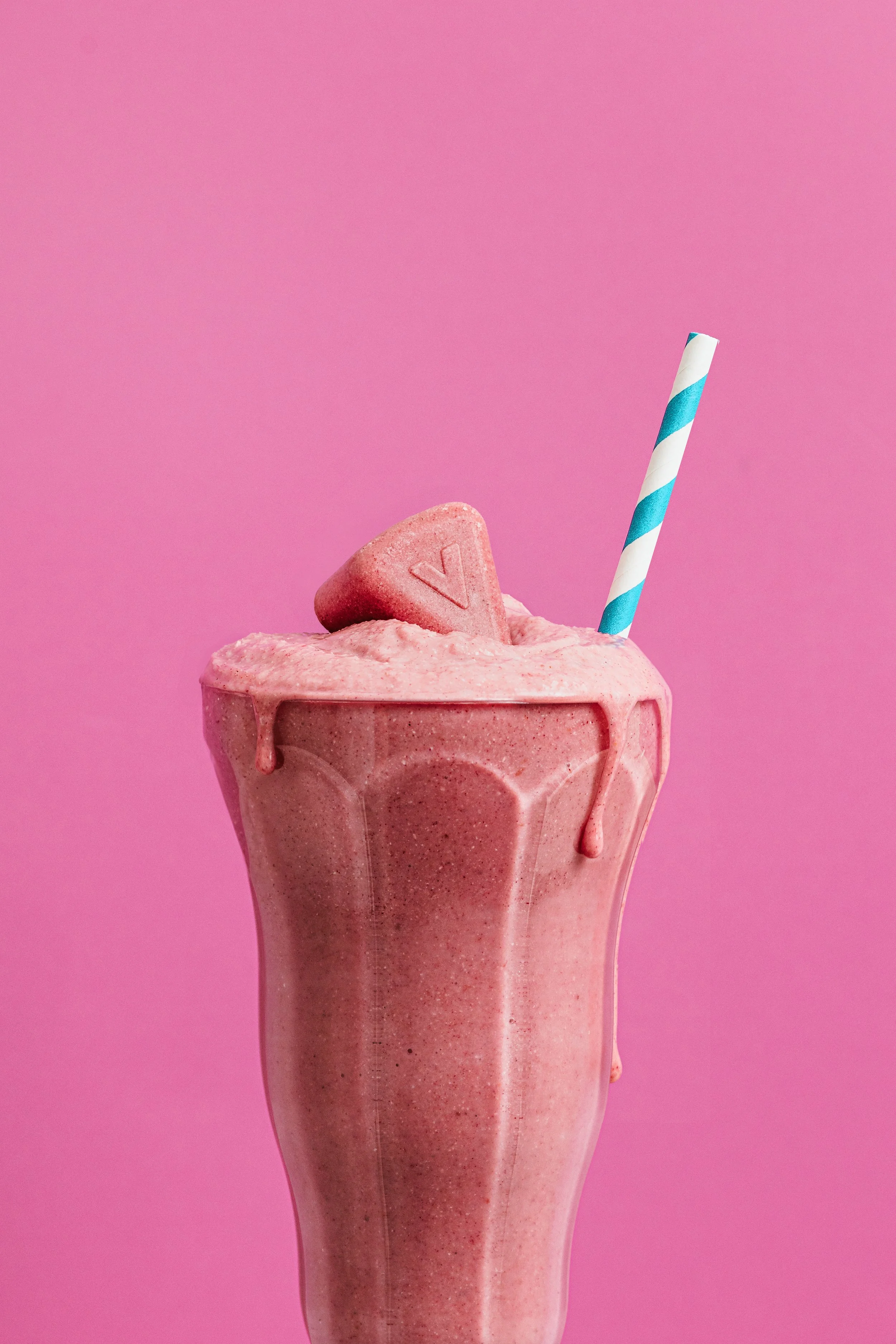 A pink milkshake topped with a pink heart-shaped candy and a striped blue and white straw against a pink background.