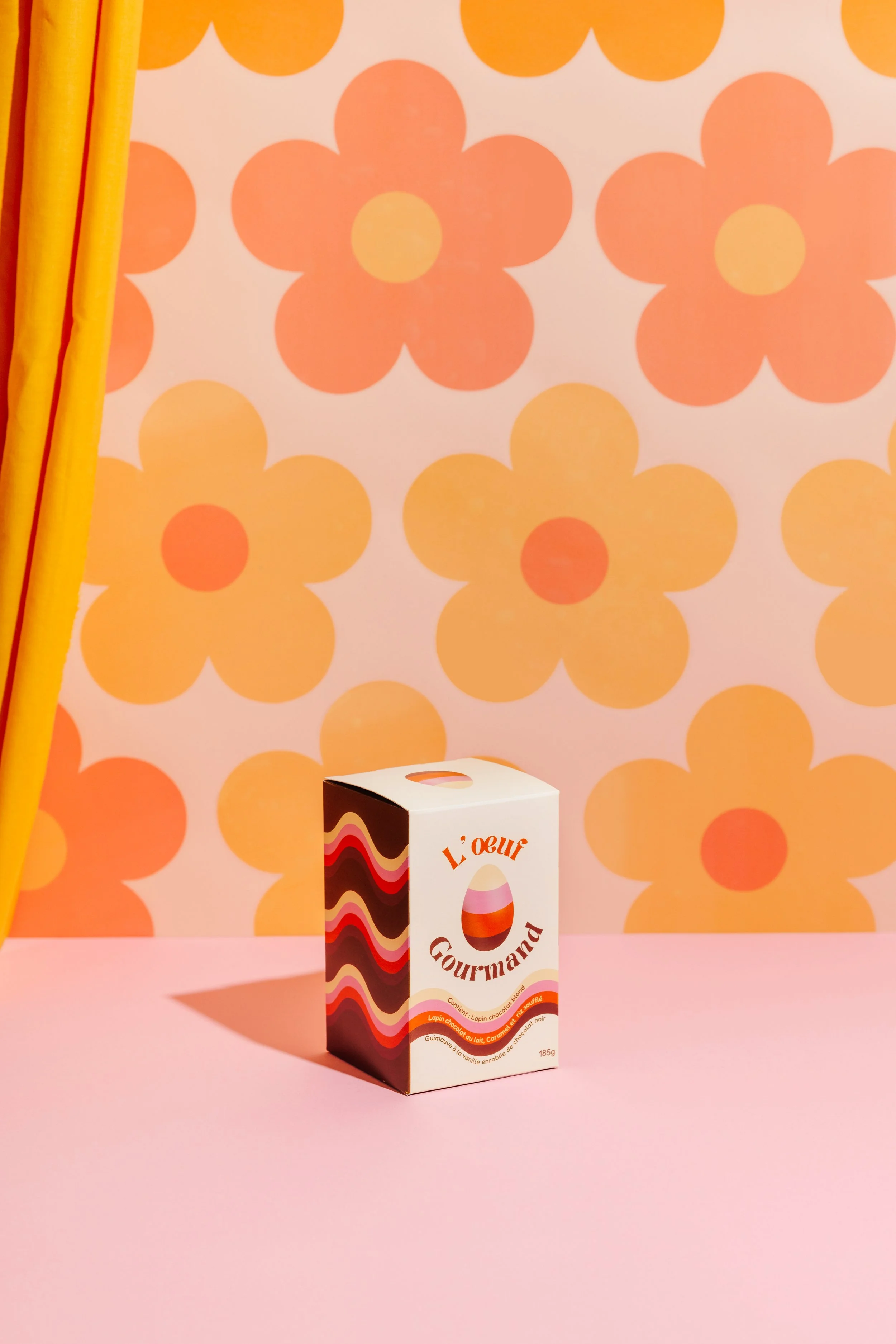 Pink table with a box of chocolate Easter eggs labeled 'L'œuf Gourmand' against a yellow and orange floral wallpaper."