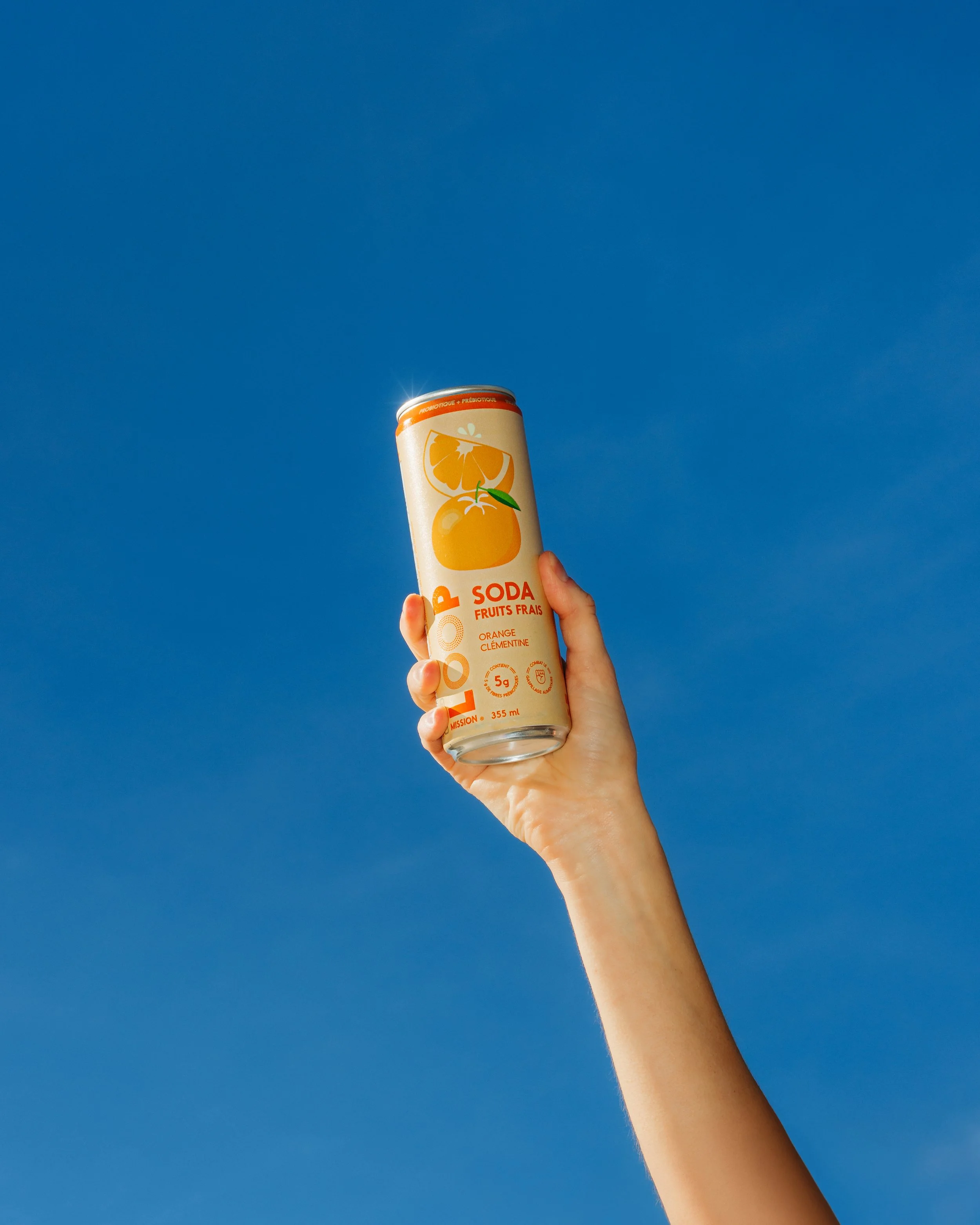 A hand holding a can of orange-flavored soda against a clear blue sky.