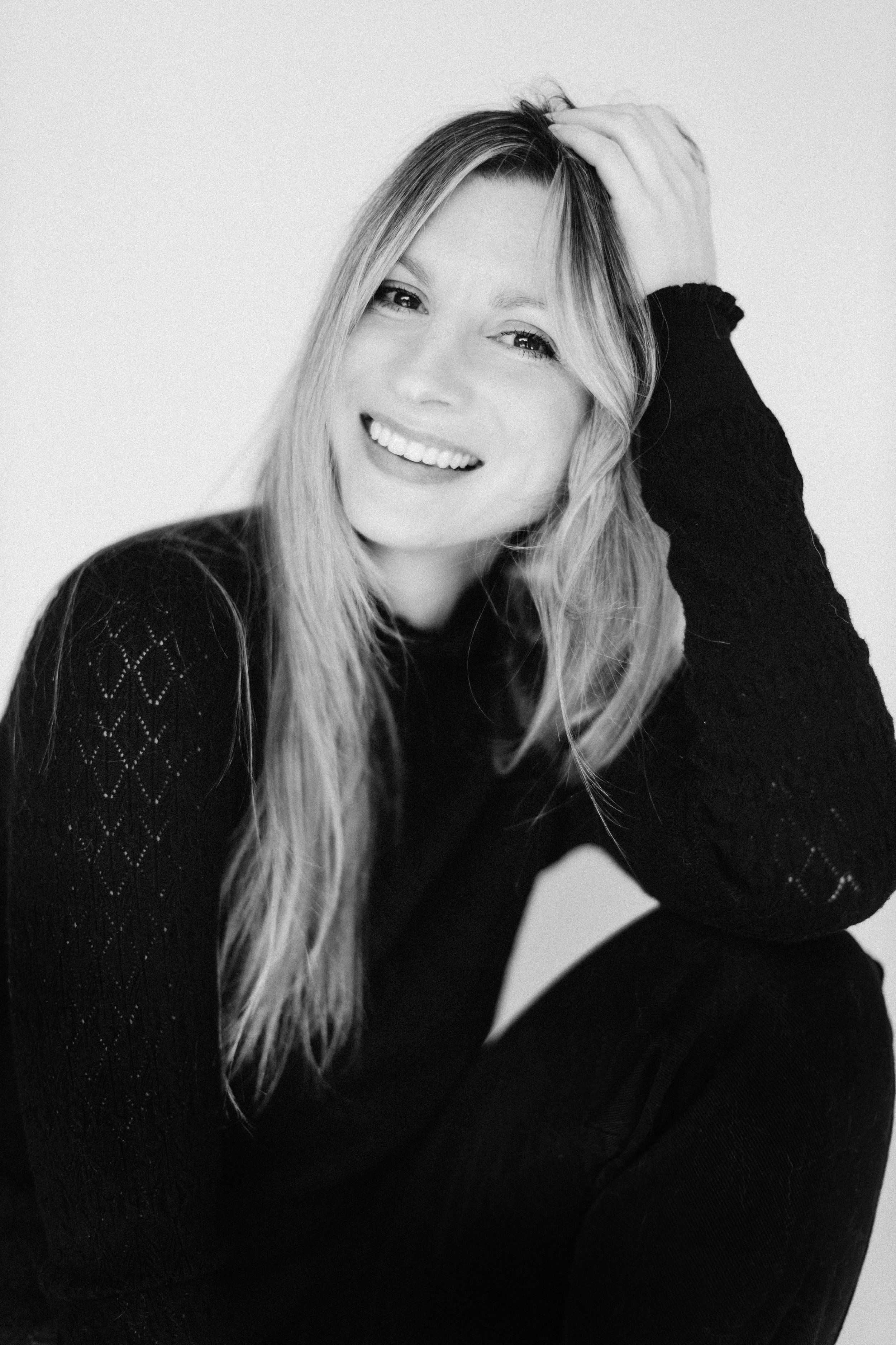 A woman with long light hair, smiling, wearing a dark sweater with a textured pattern, sitting against a plain background.