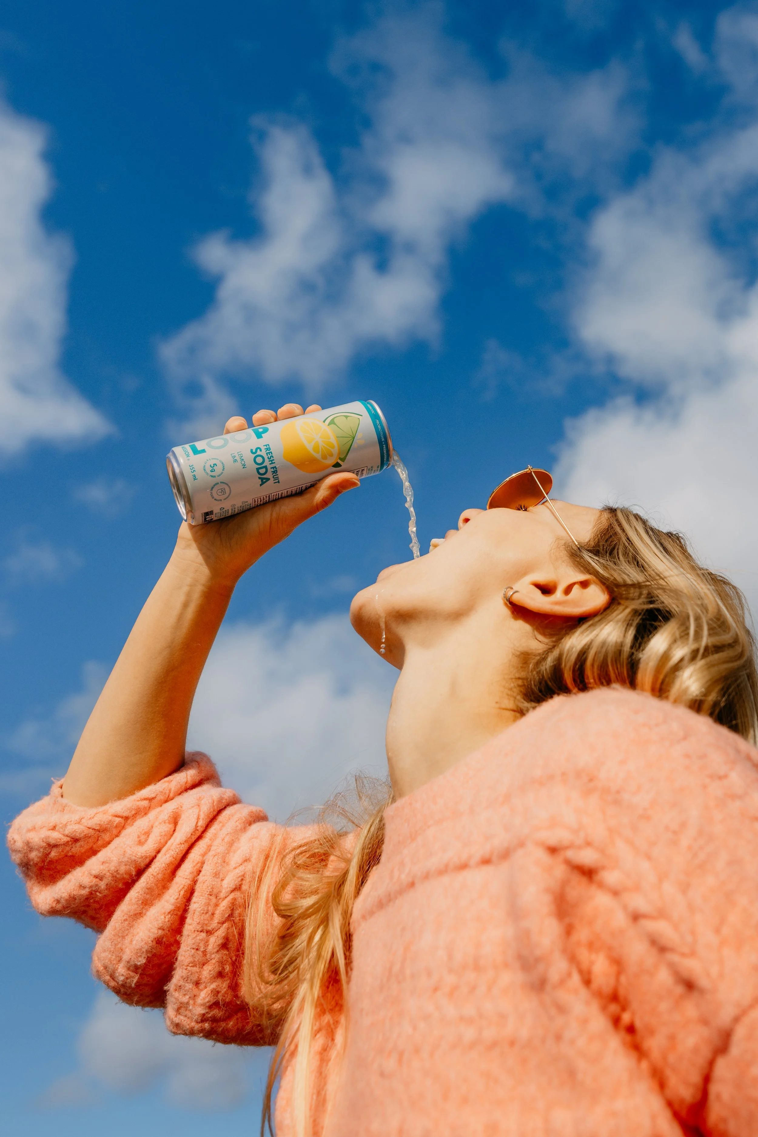 A woman with blonde hair wearing sunglasses and an orange jacket is drinking soda from a can outdoors against a sky with clouds.
