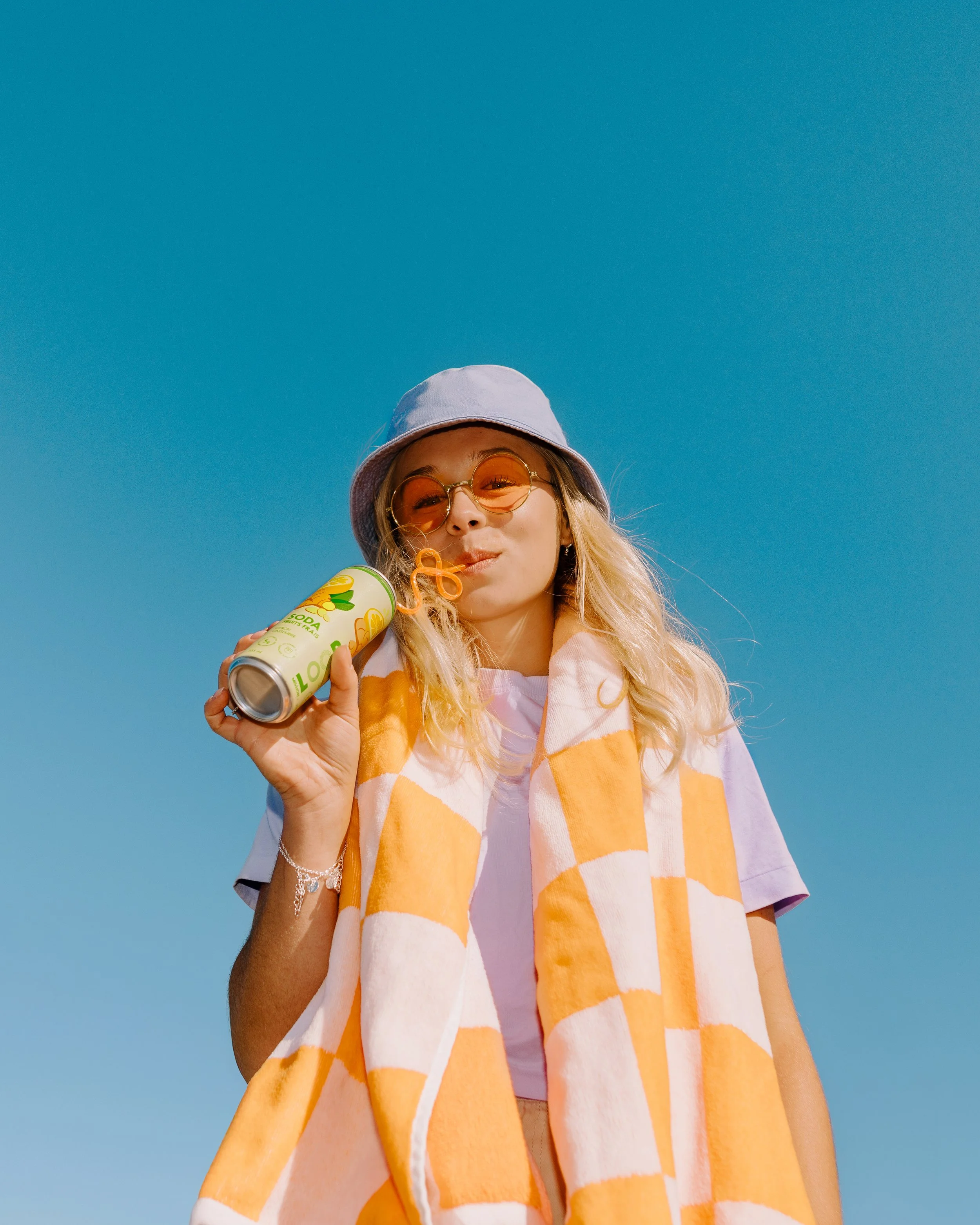 A young woman with blond hair, wearing a lavender hat and orange-tinted sunglasses, is blowing bubbles with a bubble wand while holding a canned drink with a straw. She has a yellow and orange checkered towel around her neck and is standing against a clear blue sky.