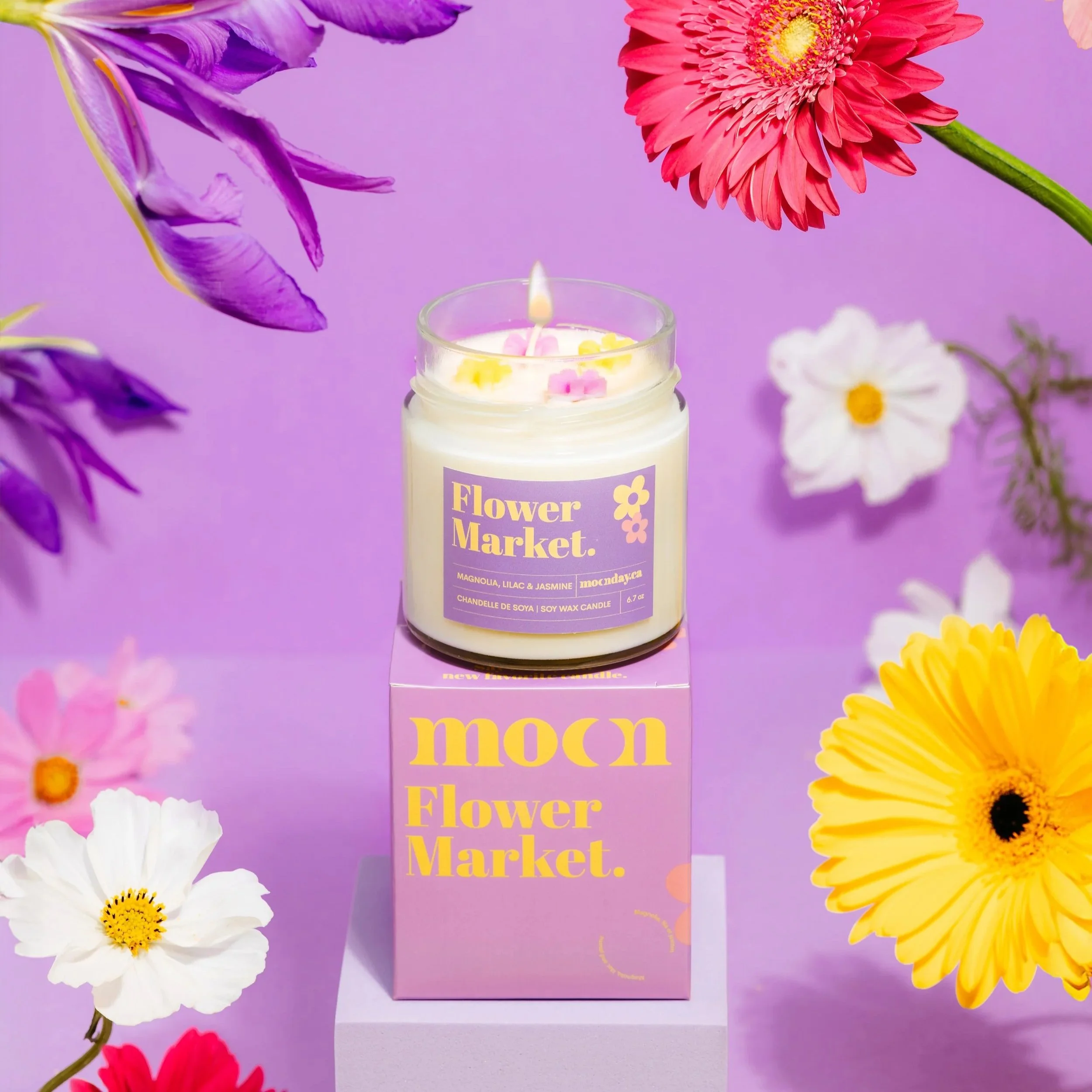 A candle with a lavender label that reads "Flower Market" is surrounded by colorful flowers including purple irises, red gerbera daisies, white daisies, and yellow daisies, against a purple background.