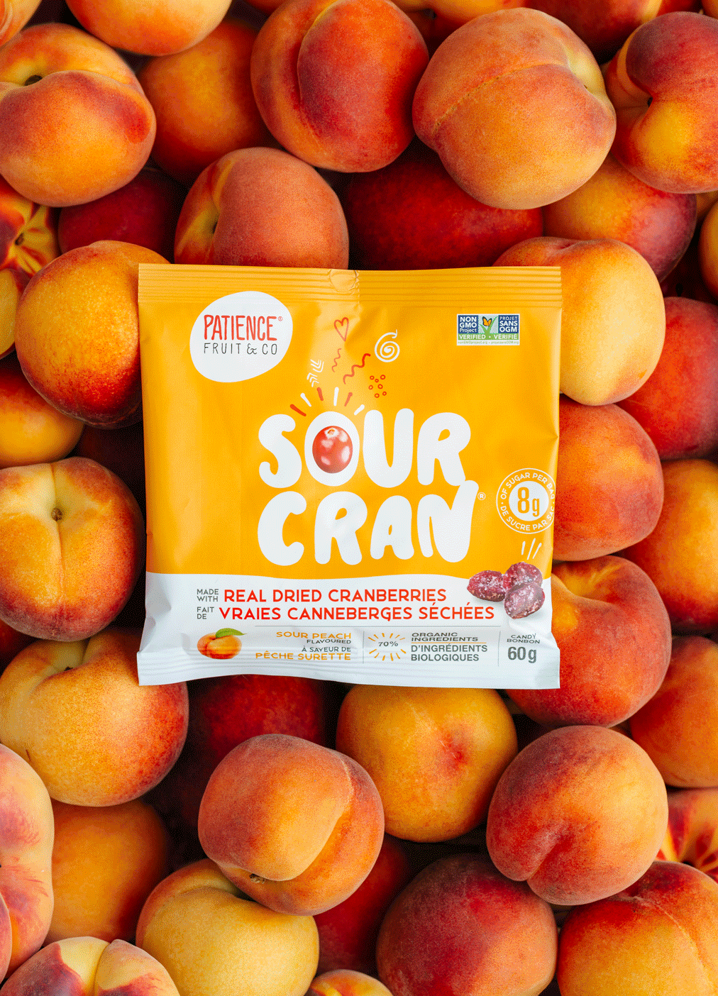 A yellow package of Sour Cran fruit chews labeled 'Patience Fruit & Co' surrounded by fresh peaches and nectarines.