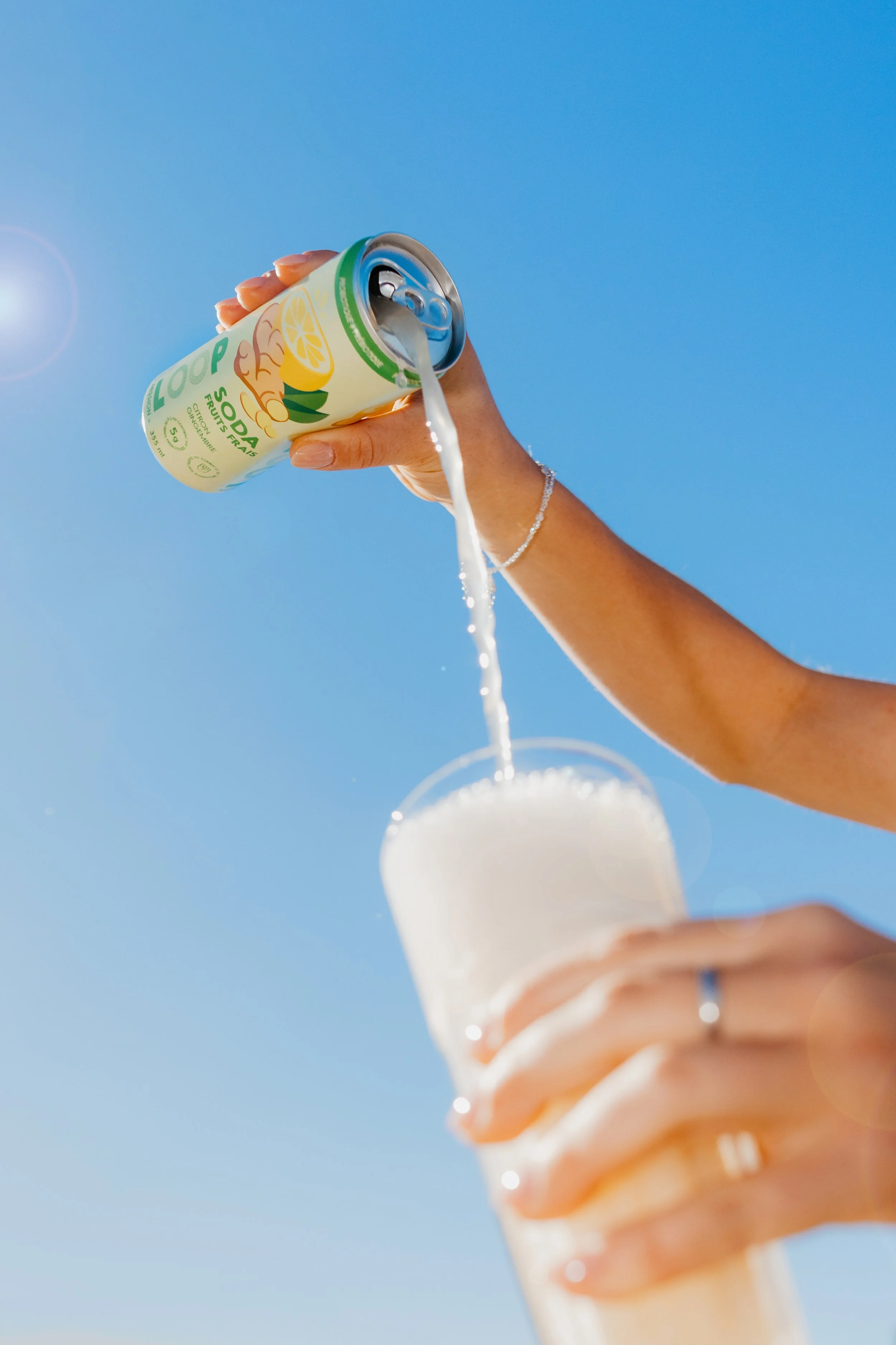 Person pouring a can of citrus-flavored soda into a glass against a clear blue sky.