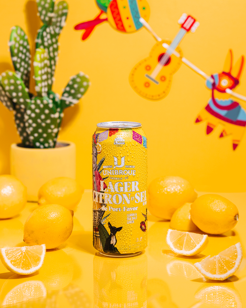 A can of Unibroue lager with lemon flavor on a yellow background, surrounded by whole and sliced lemons, with a cactus plant in a yellow pot on the left and colorful decorative wall art featuring a guitar, sun, and animal on the yellow wall.