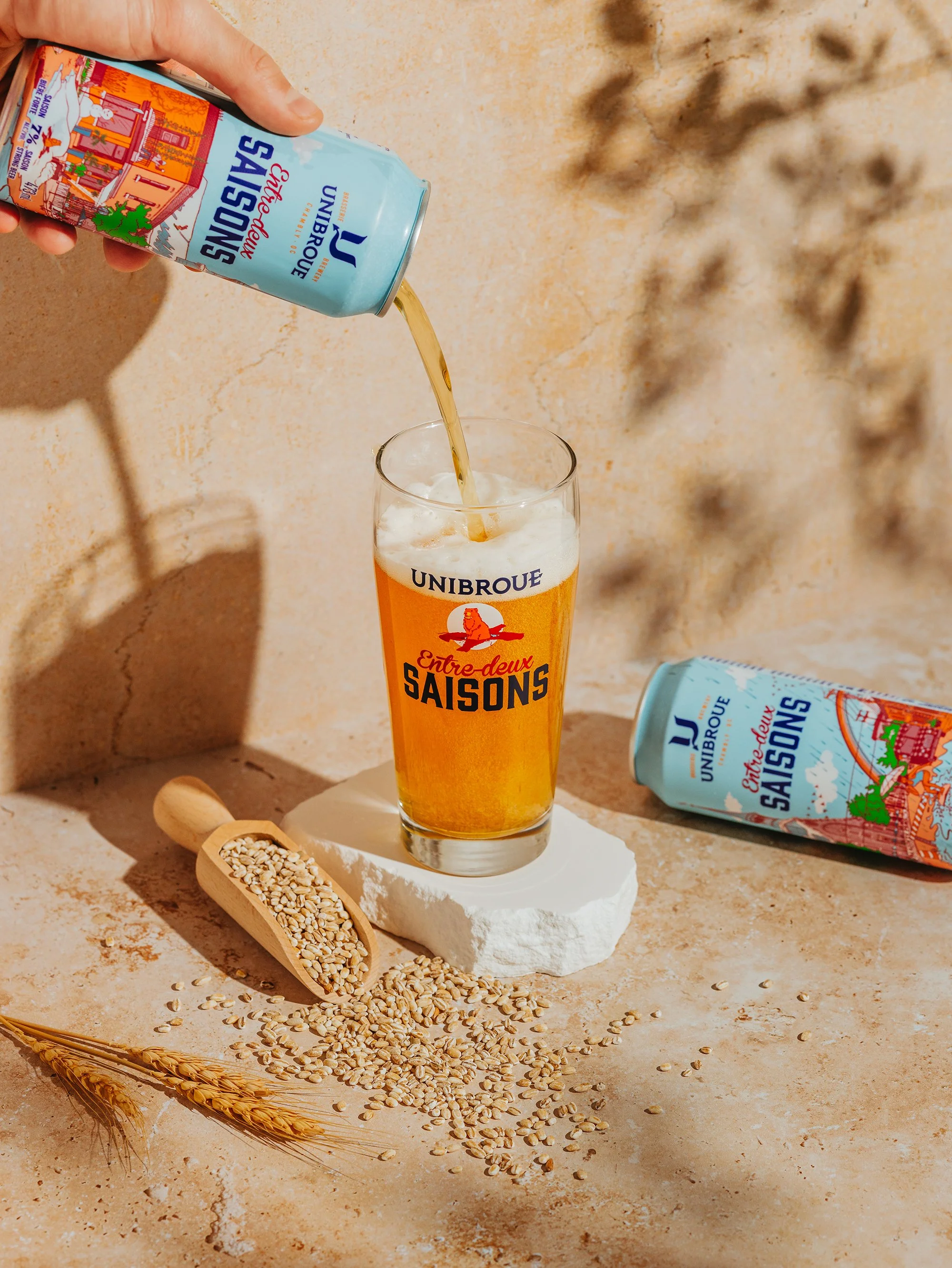 A hand pouring sparkling water from a can labeled 'Eau Minérale Naturelle Gazeuse' into a glass with 'Unibroue' logo, with a scoop of oats and wheat stalks on a marble surface.
