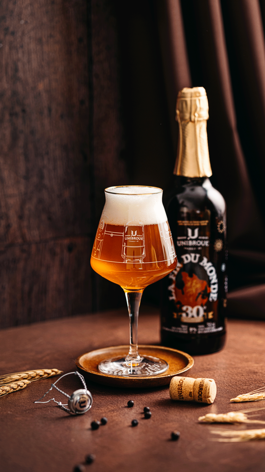 A glass of amber beer with a foamy head on a wooden surface, with a dark bottle of beer in the background, a cork, black peppercorns, and wheat stalks around it.