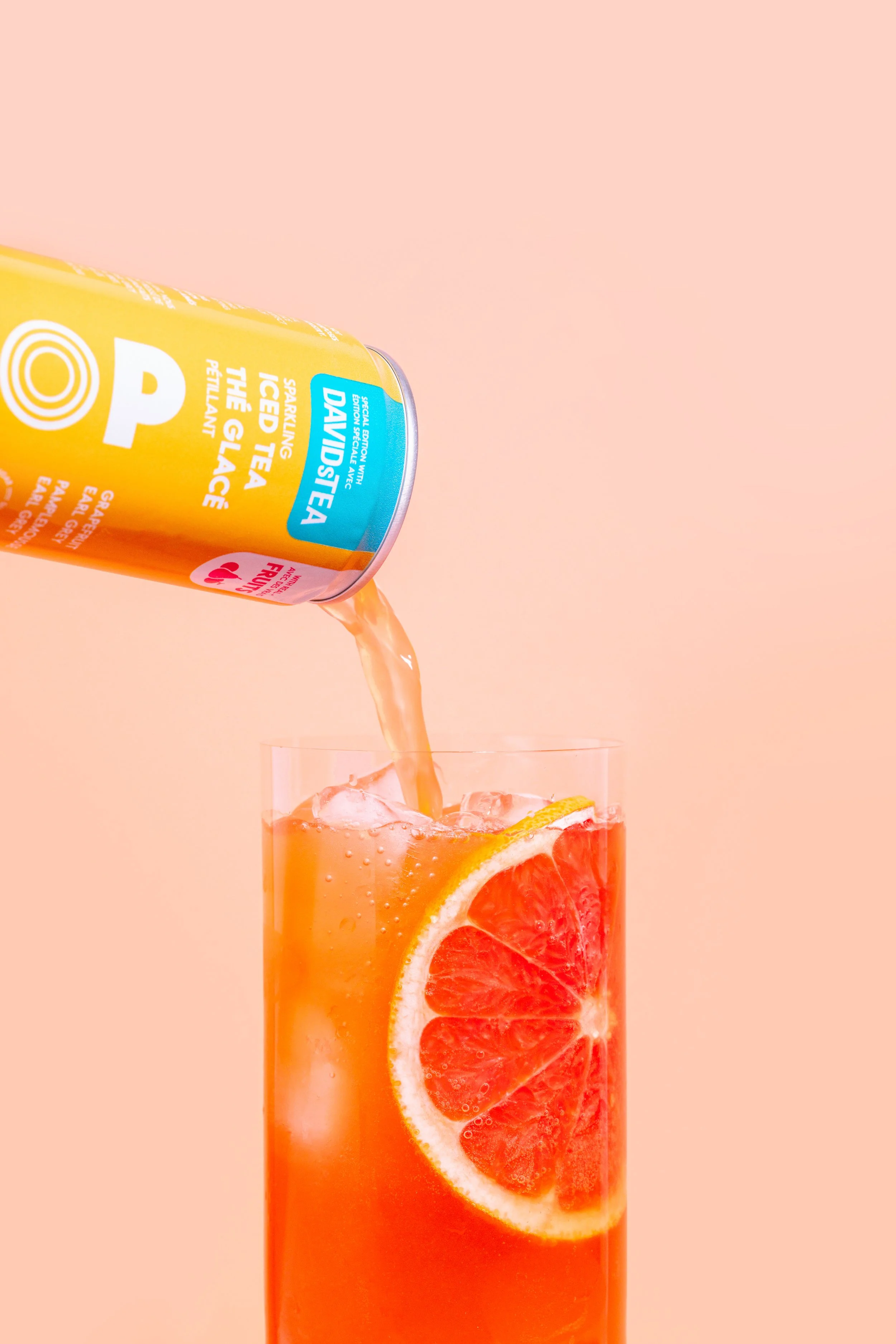 A can of Davidstea Sparkling Iced Tea The Glacé pouring over a glass filled with ice and grapefruit slices, with a pink background.