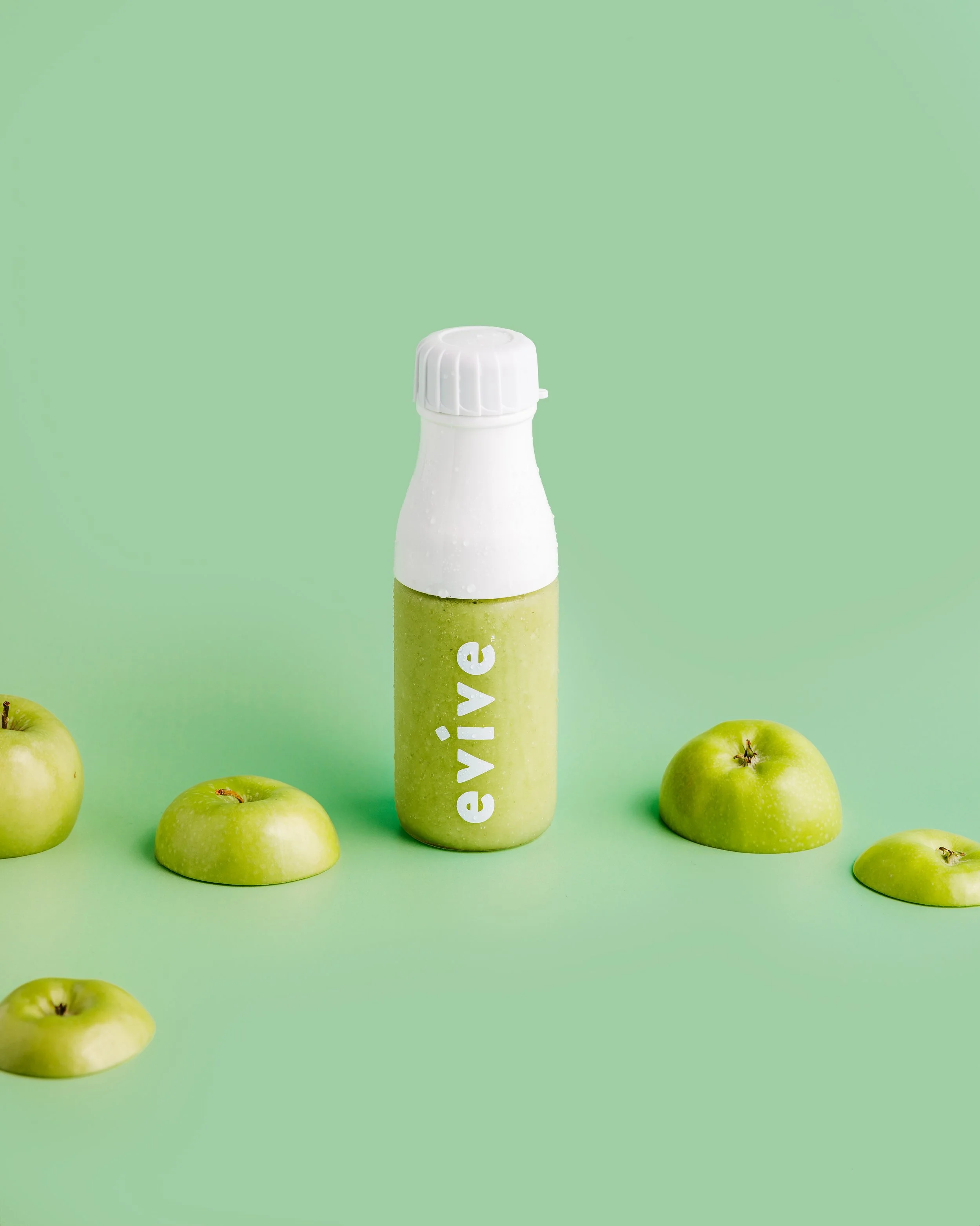 A small bottle with a white cap and green body that has the word "e-vive" written on it, surrounded by several green apples on a light green background.
