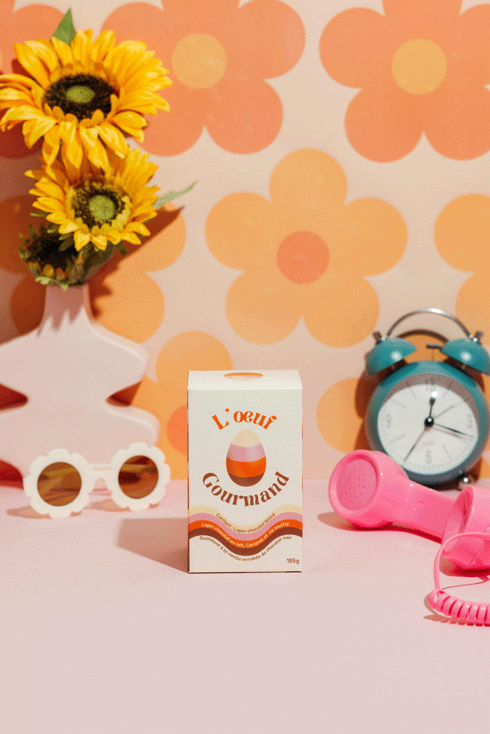 A pink surface with a box of chocolate labeled "L’œuf Gourmand" in the center, a vintage teal alarm clock, a pink telephone handset, a white decorative object shaped like a pair of glasses, and a small white vase with yellow sunflowers, with a background of large orange flower patterns.