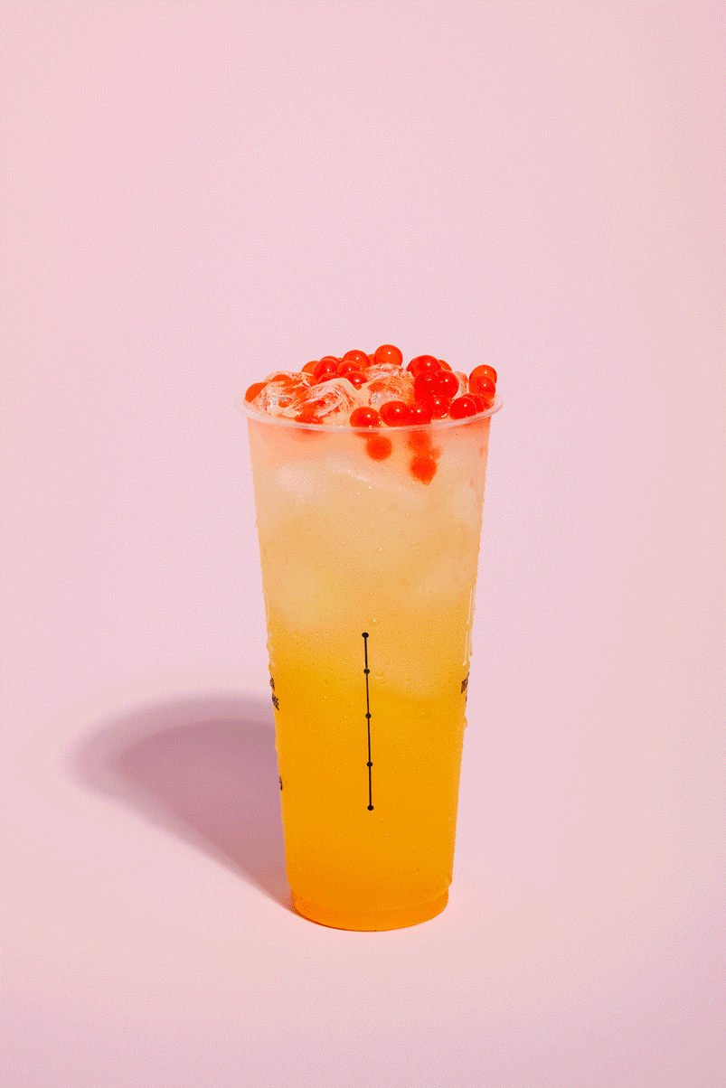 A tall clear plastic cup filled with a yellow-orange beverage, topped with ice and red tapioca pearls, against a light pink background.