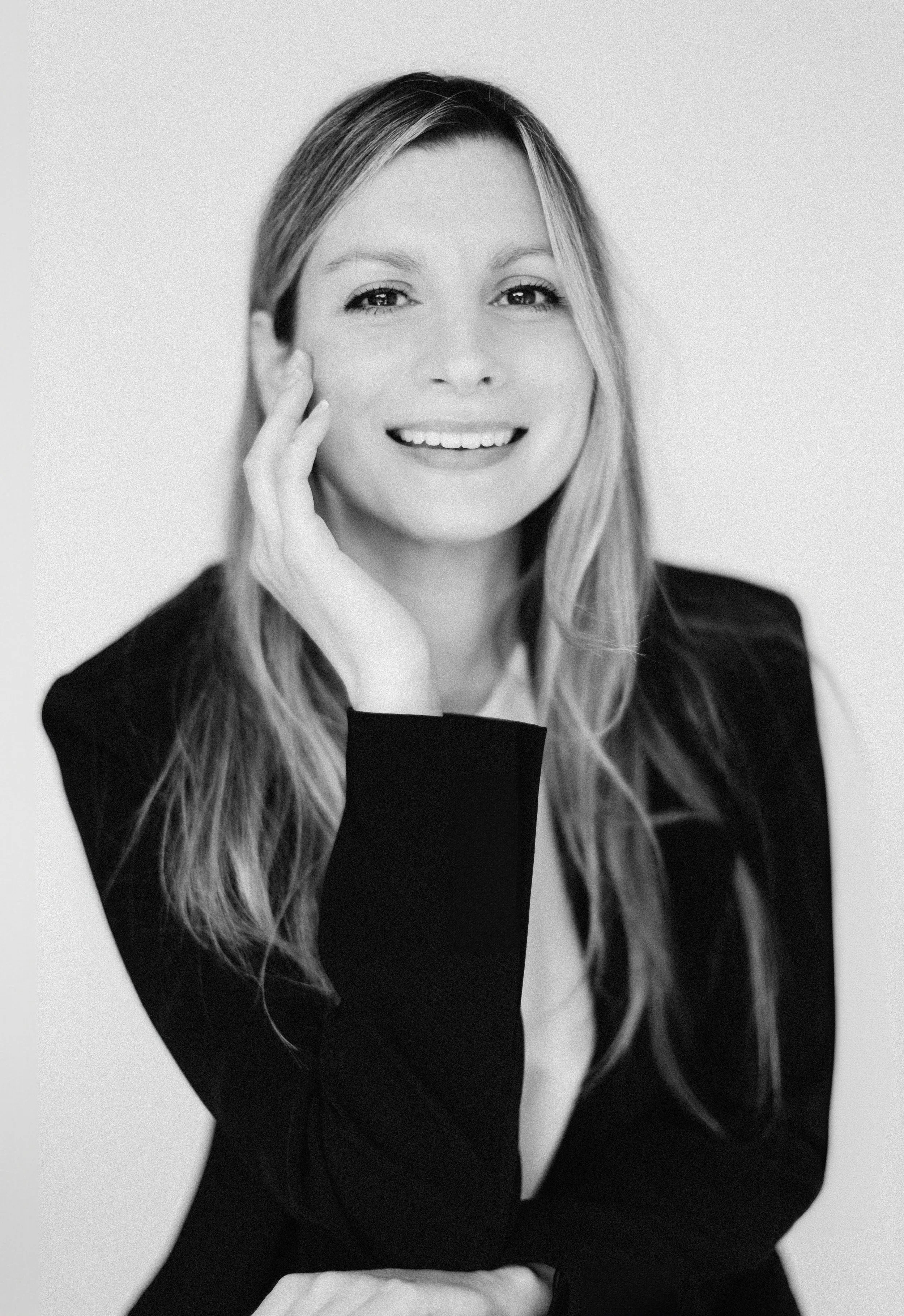 Black and white portrait of a woman with long hair smiling, touching her face, wearing a dark blazer.