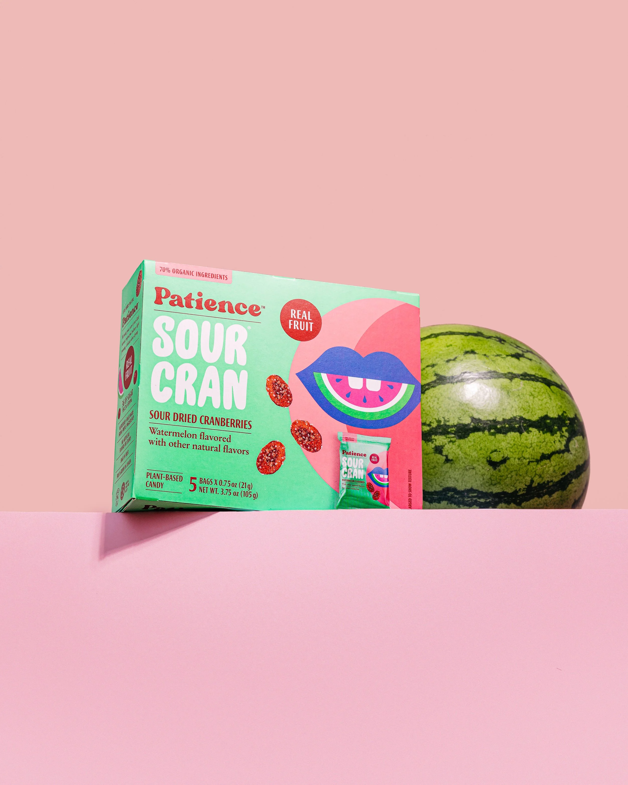 A box of Patience sour cranberry candies with a watermelon flavor, placed on a pink surface next to a watermelon.