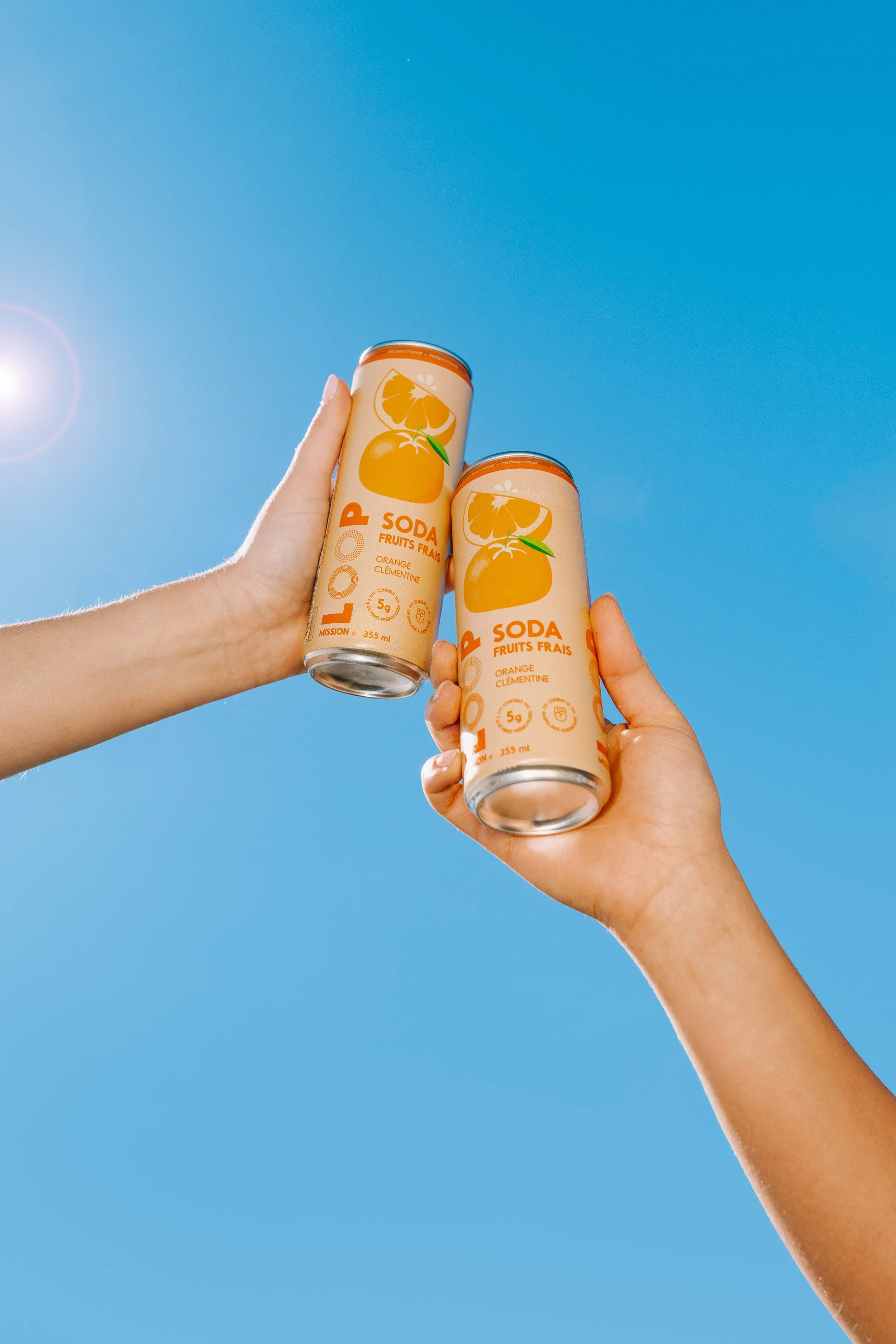 Two hands holding cans of orange flavored soda against a clear blue sky.